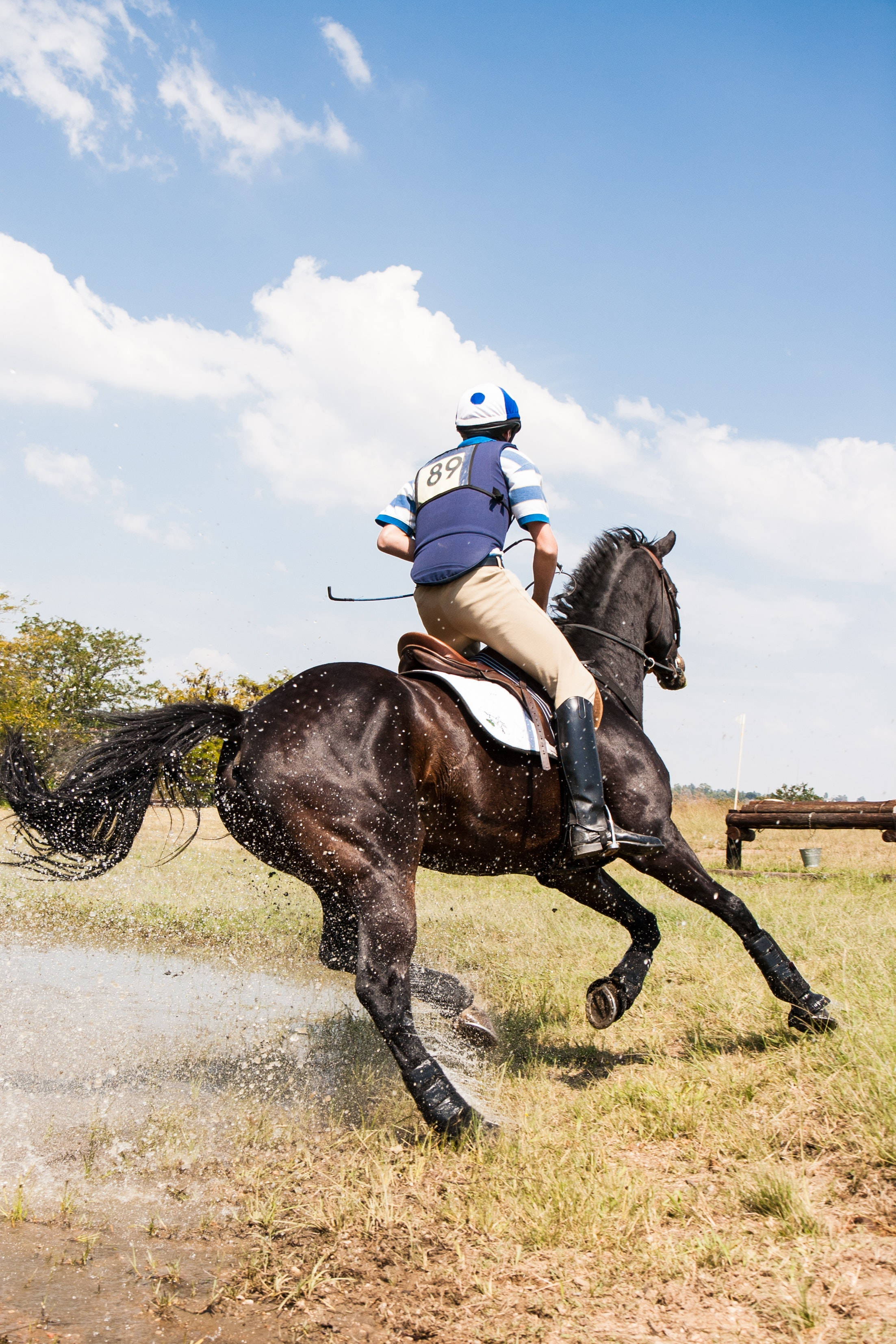 Horse iPhone 桌布 壁紙 — 一名身穿藍色背心和頭盔的騎手，在晴朗藍天和蓬鬆白雲下，騎著黑馬飛奔過泥濘的水坑。