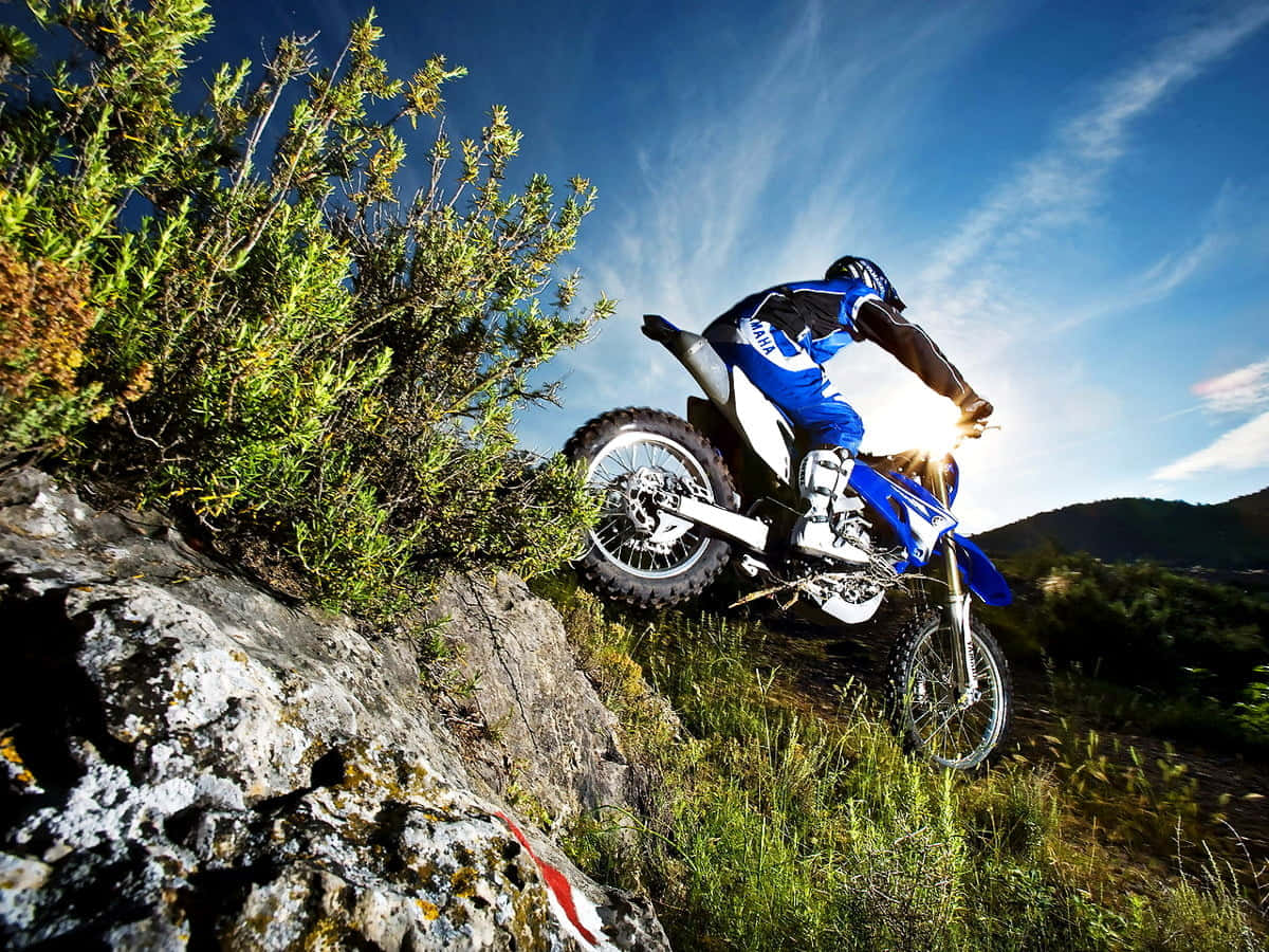 Motocross 桌布 壁紙 — 一名身穿藍色裝備的越野電單車手，在陽光耀眼的明亮藍天下，於岩石小徑上傾身過急彎。