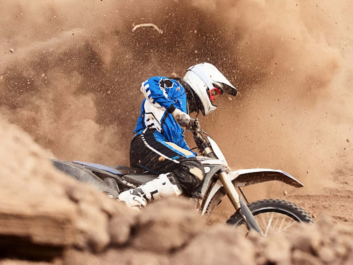 Dirtbike 桌布 壁紙 — 一名身穿藍色裝備的越野電單車手在崎嶇的泥地上轉彎，揚起大片棕色塵土。