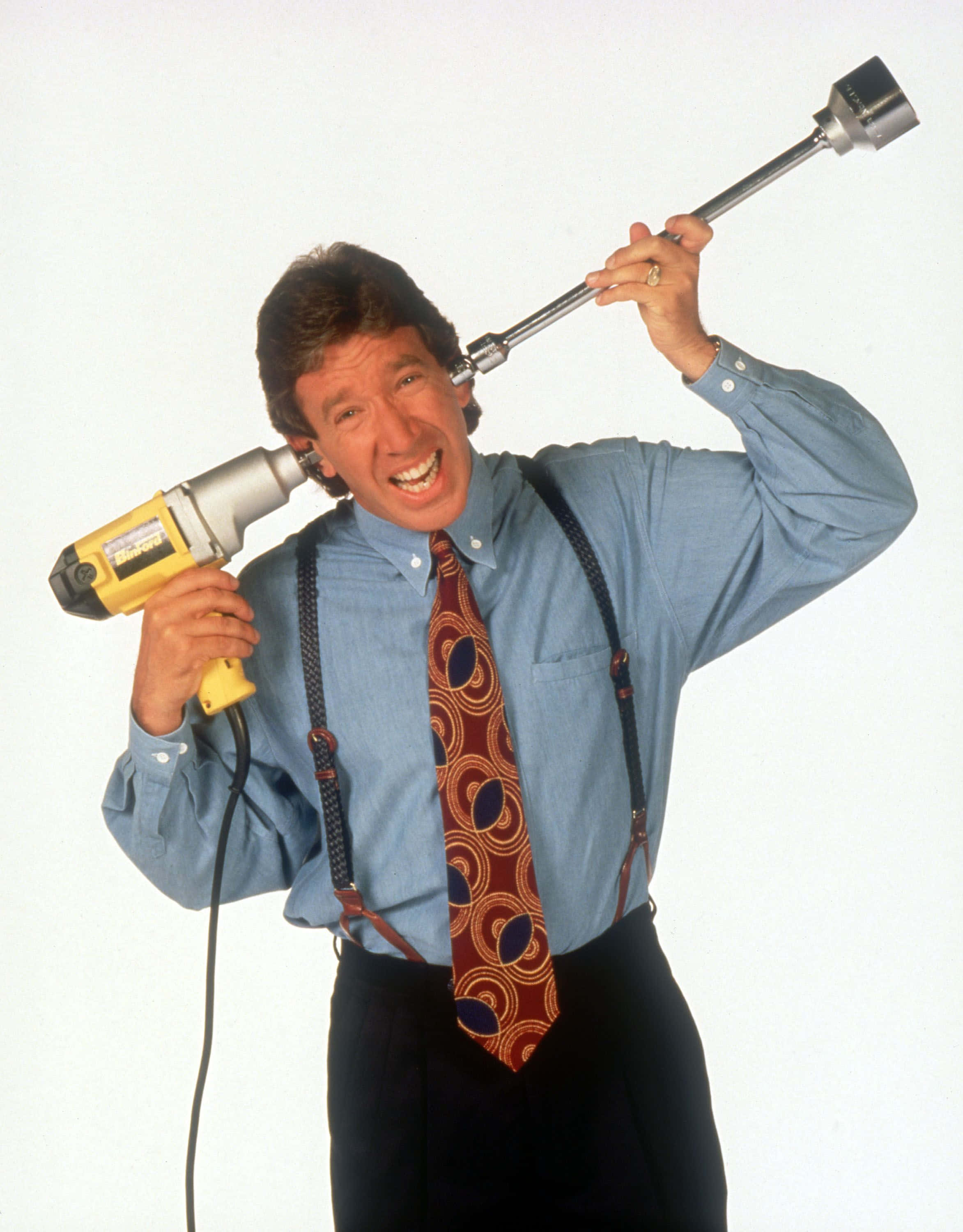 Tim Allen 桌布 壁紙 — 一名身穿藍色襯衫和圖案領帶的男子，在純白色背景前，將黃色電鑽像咪高峰一樣舉起，咧嘴而笑。