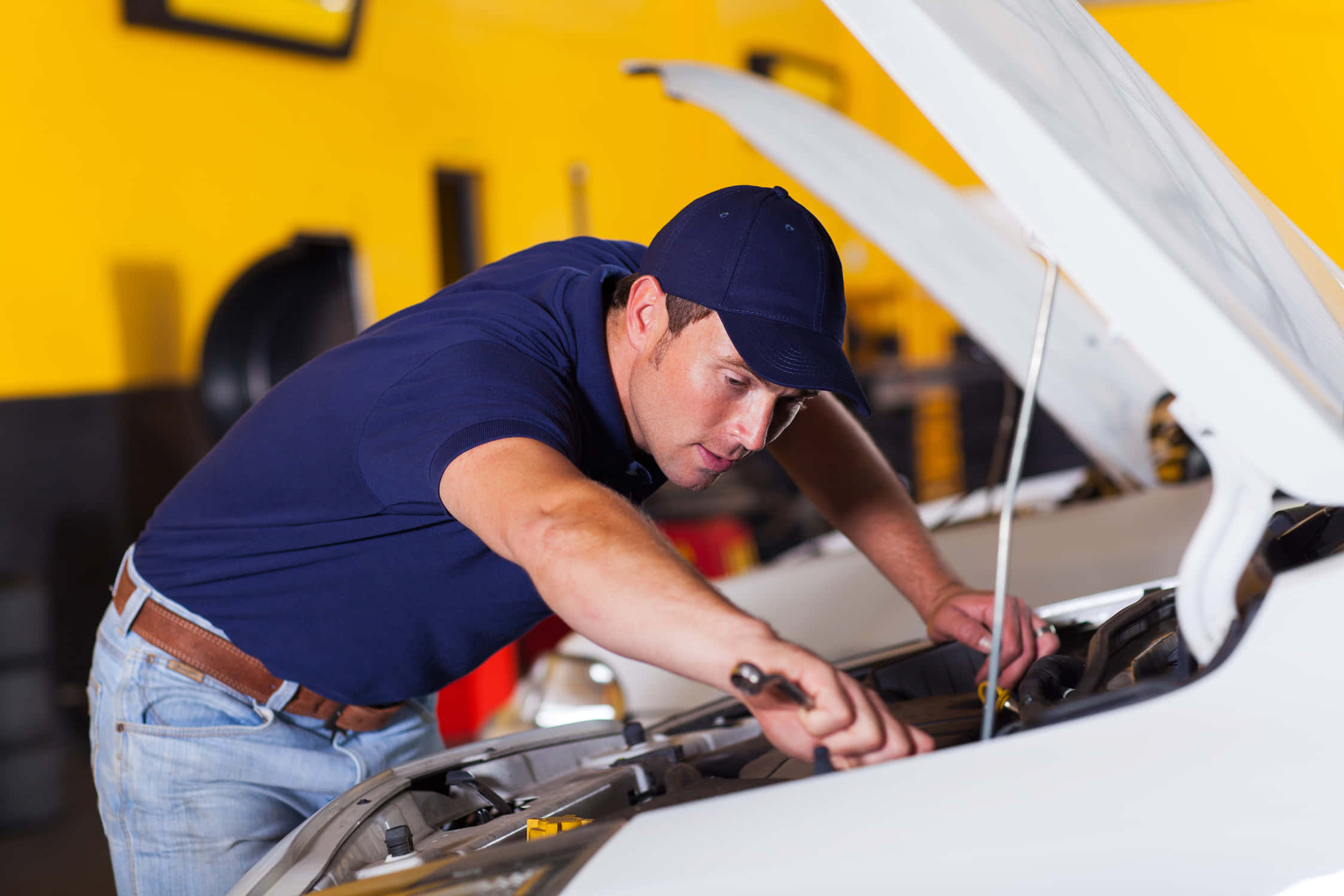 Car Repair 桌布 壁紙 — 一名身穿藍色襯衫和帽子的機械師，在明亮的黃色車庫裡，俯身於一輛白色汽車的打開引擎蓋上，使用扳手。