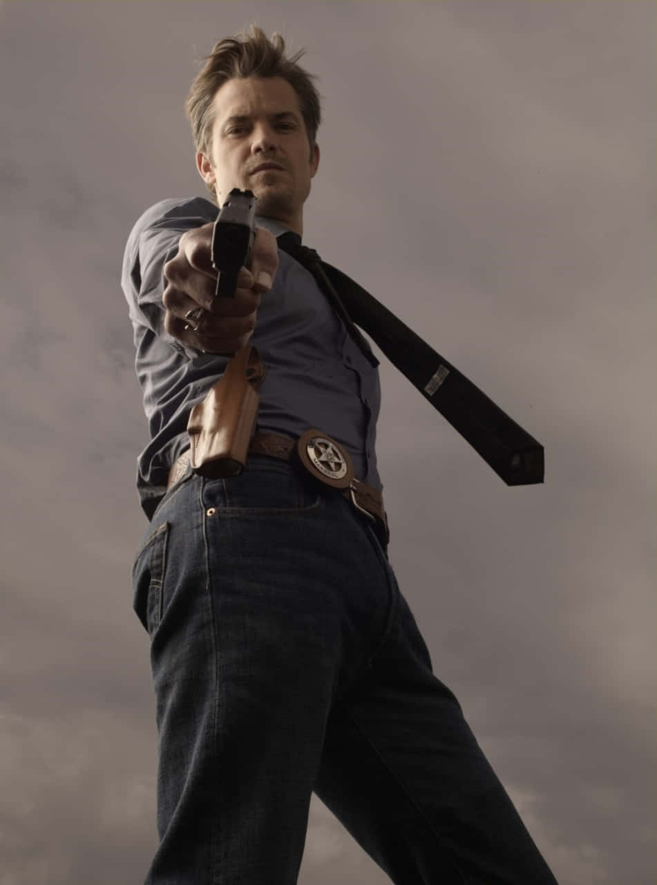 timothy olyphant 桌布 壁紙 — 一名身穿藍色襯衫和牛仔褲的男子，在陰雲密布的天空下，直接將手槍瞄準觀眾。
