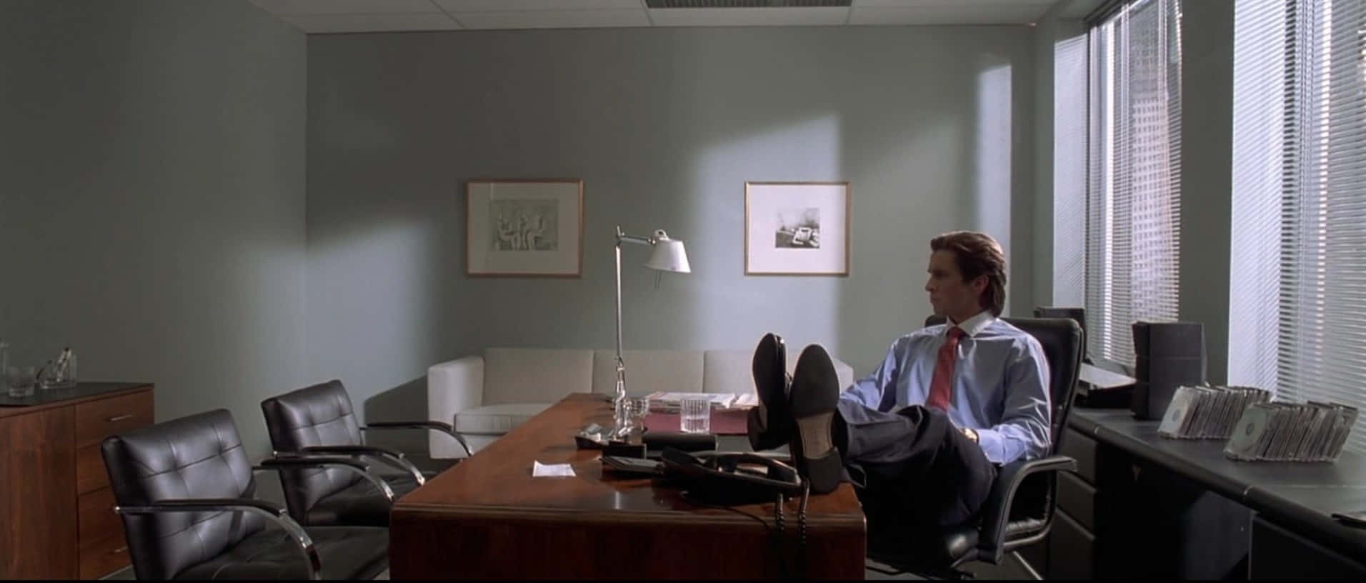 patrick bateman 桌布 壁紙 — 一名身穿藍色襯衫和紅色領帶的男子，在一個充滿柔和日光的簡約辦公室裡，將腳擱在木桌上放鬆。