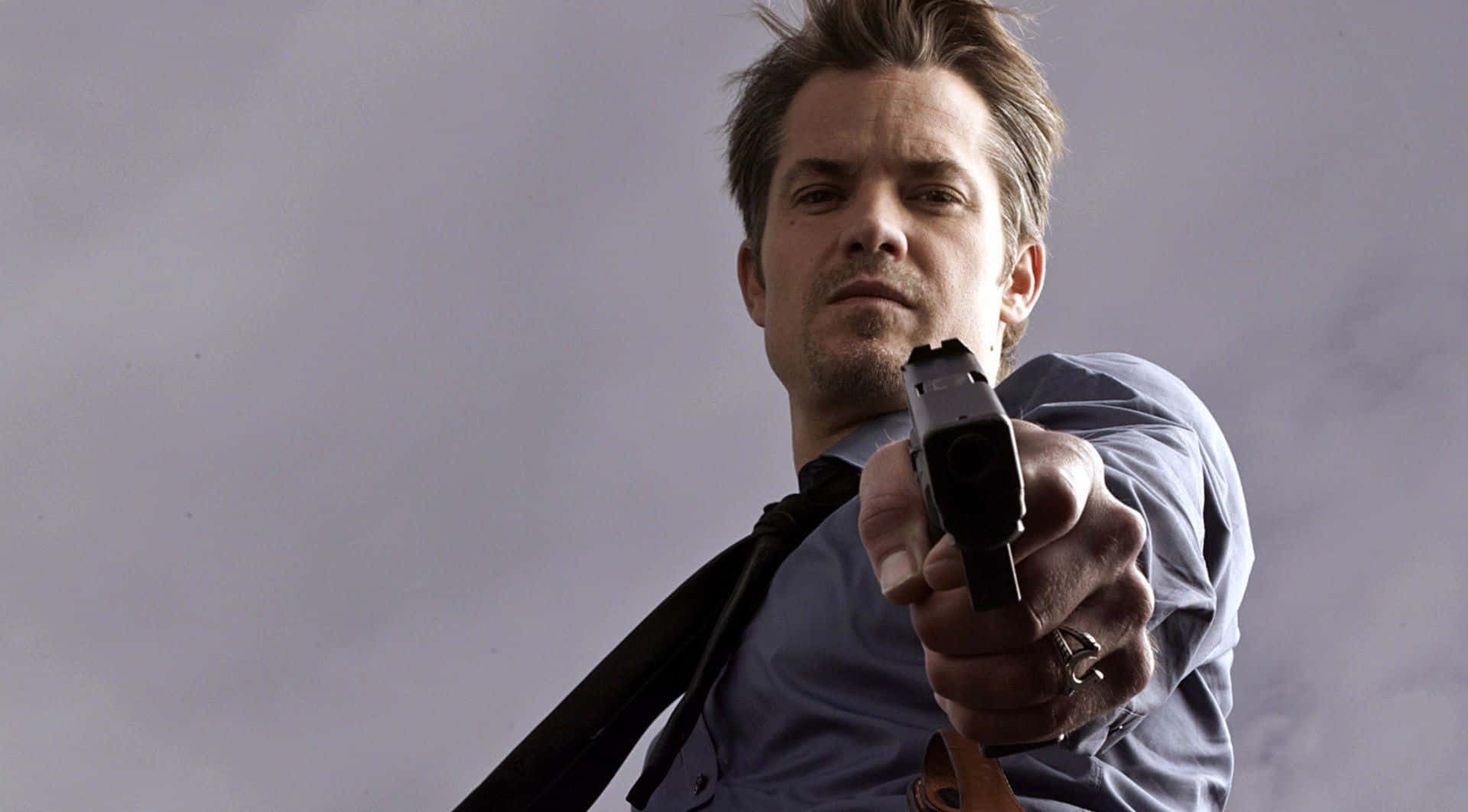 timothy olyphant 桌布 壁紙 — 一名身穿藍色襯衫和領帶的男子，在灰色天空下，將手槍直接瞄準觀眾。