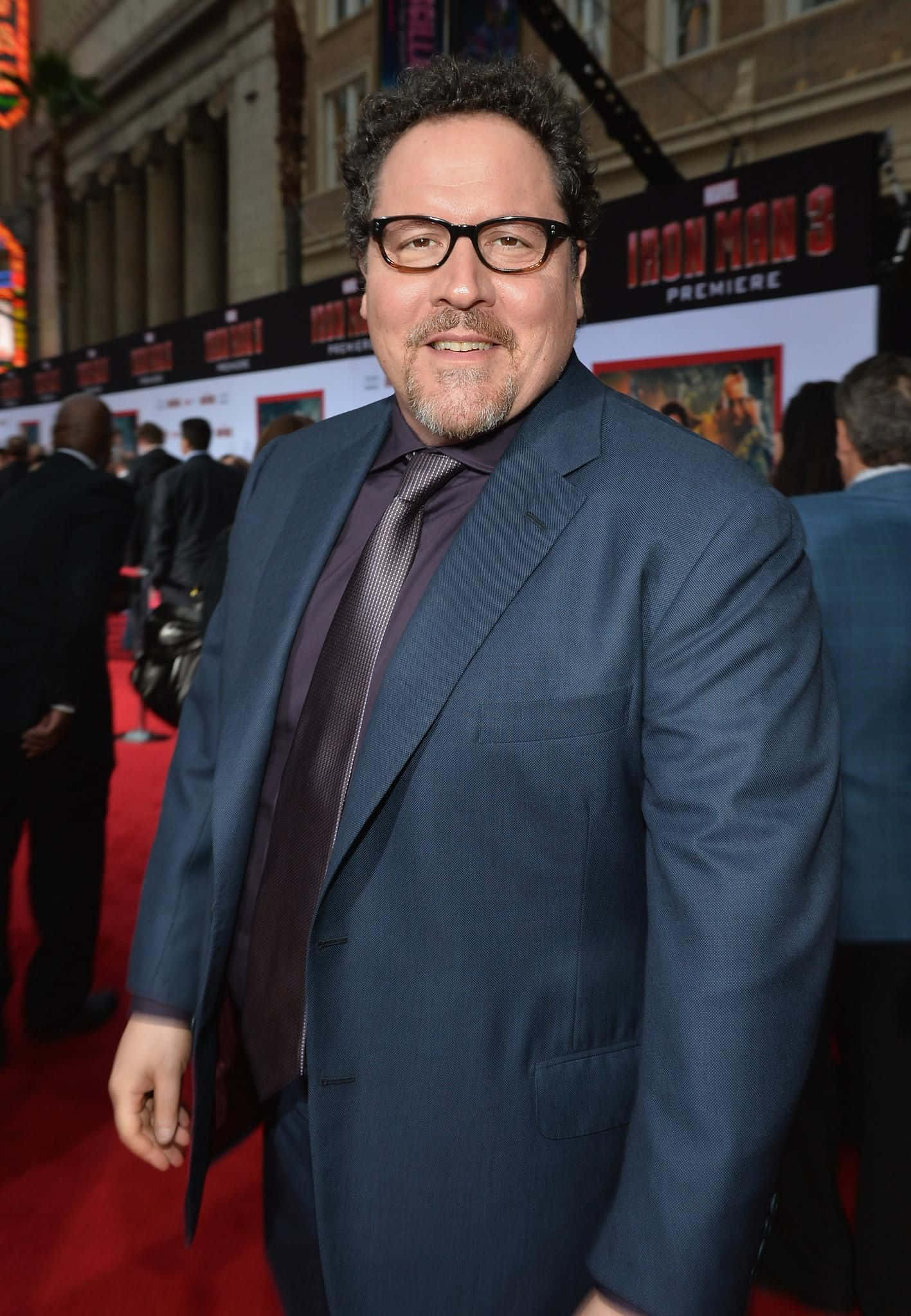 Jon Favreau 桌布 壁紙 — 一名身穿藍色西裝及戴眼鏡的男子在《鐵甲奇俠3》首映禮的紅地毯上微笑，身後有電影海報。