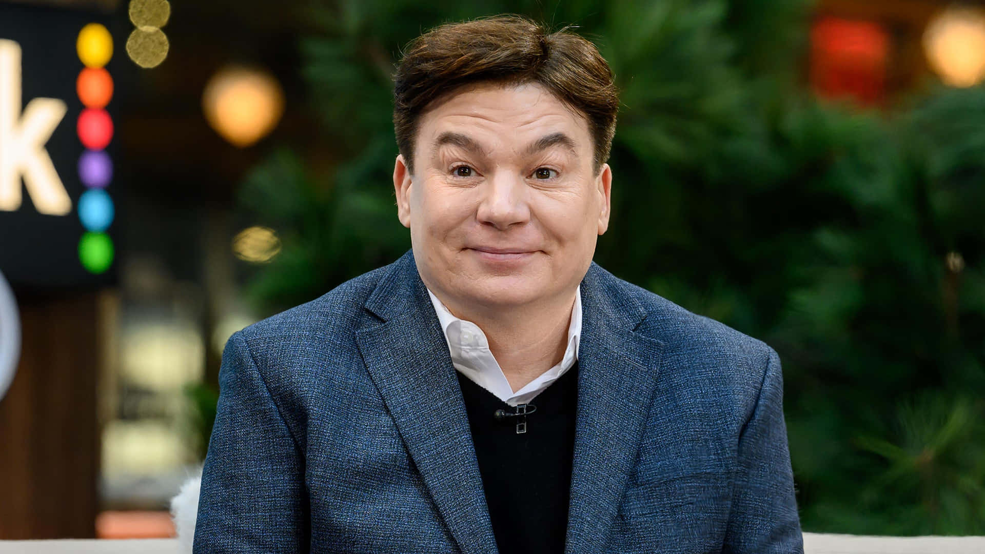 Mike Myers 桌布 壁紙 — 一名身穿藍色西裝外套的男子，在色彩繽紛燈光下柔和模糊的節日背景前，對著鏡頭溫暖微笑。