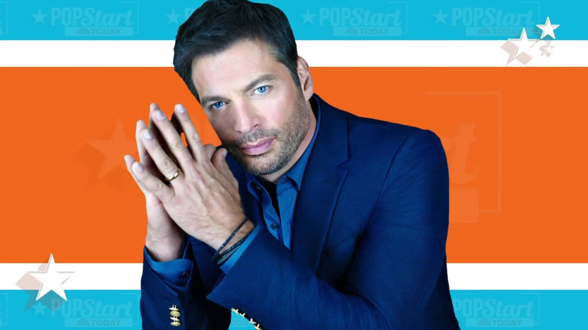 Harry Connick Jr 桌布 壁紙 — 一名身穿藍色西裝的男子，在帶有星星圖案的鮮明橙色和藍綠色條紋背景前，雙手合十擺姿勢。