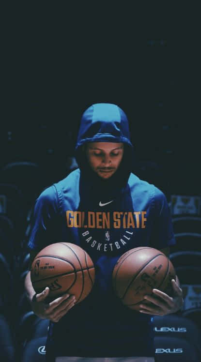 steph curry 桌布 壁紙 — 一名身穿藍色連帽衫的籃球運動員在昏暗的場館燈光下，專注地拿著兩個籃球。