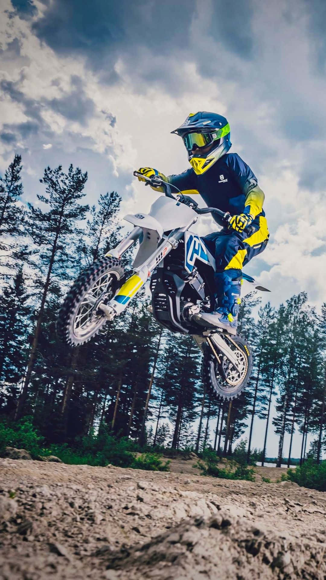 Dirtbike 桌布 壁紙 — 一名身穿藍黃色裝備的越野電單車手，在陰天之下，騎著白色越野電單車飛越森林小徑上空。