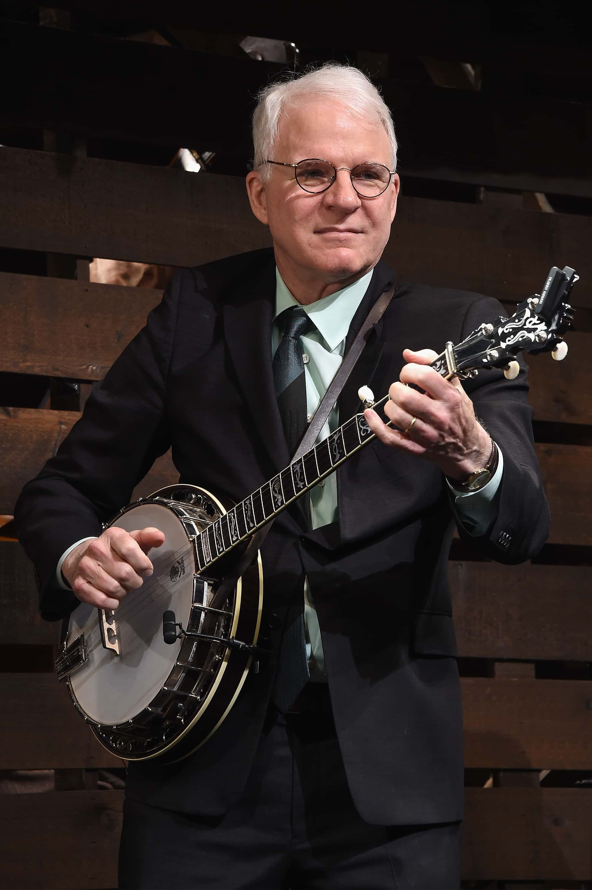 steve martin 桌布 壁紙 — 一名身穿西裝、戴眼鏡的男子，在深色木條背景前拿著班卓琴。