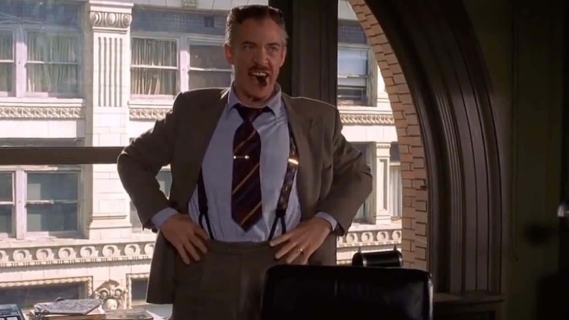 J Jonah Jameson 桌布 壁紙 — 一名身穿西裝和吊帶的男人自信地站在辦公室，雙手叉腰，張嘴大喊，陽光從他身後的大窗戶射進來。