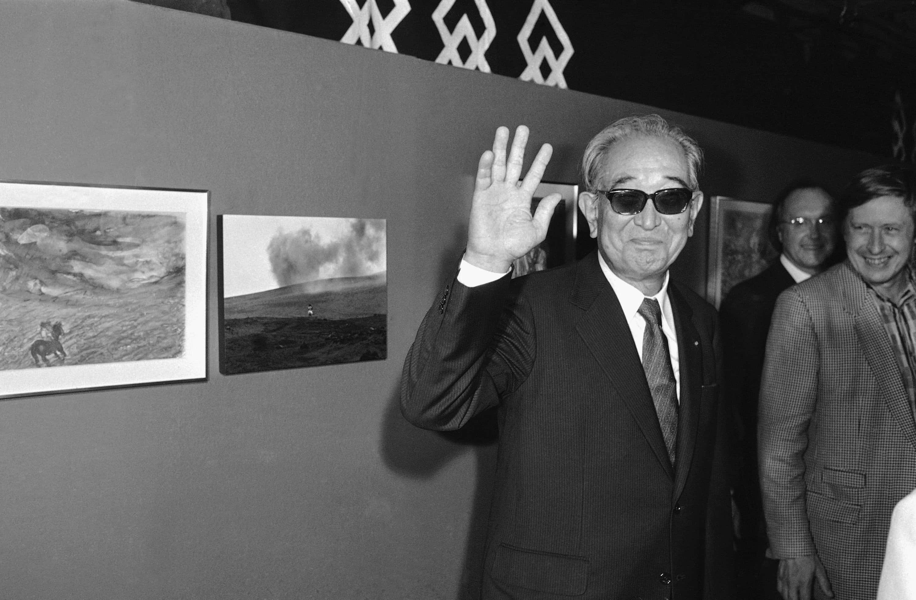 Akira Kurosawa 桌布 壁紙 — 一名身穿西裝和太陽眼鏡的男子在畫廊開幕式上揮手，身後牆上掛著裱框的風景照片。