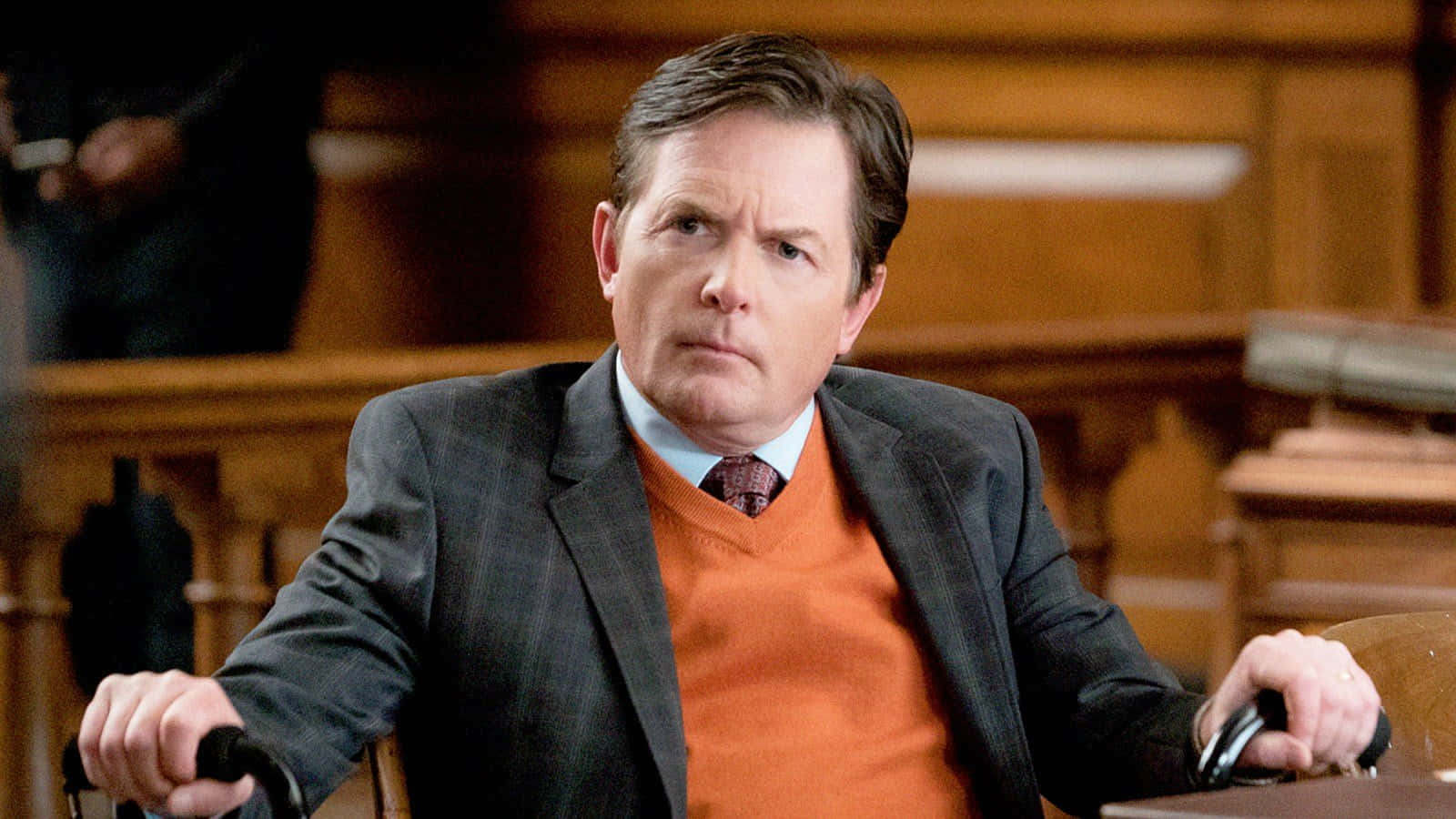 michael j fox 桌布 壁紙 — 一名身穿西裝和橙色毛衣的男子坐在法庭椅子上，表情嚴肅地抓緊扶手。