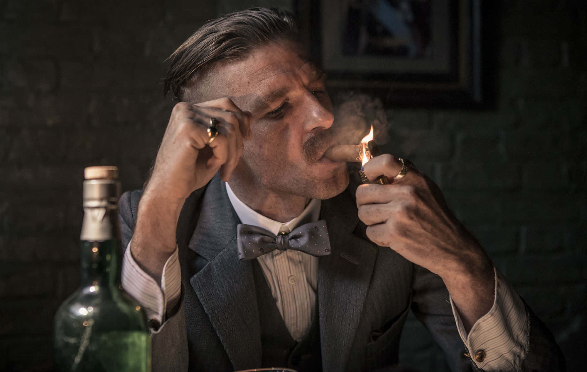 Peaky Blinders 桌布 壁紙 — 一名身穿西裝和領結的男子在光線昏暗的磚牆房間裏點燃雪茄，旁邊放著一個瓶子。