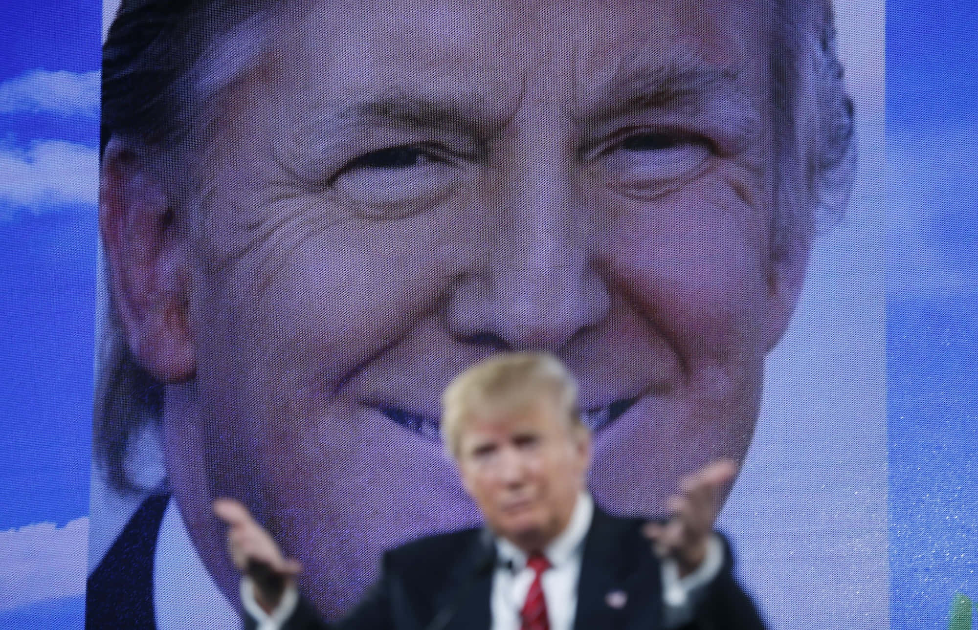 trump 桌布 壁紙 — 一名身穿西裝的男子在一個顯示他自己微笑臉孔的巨型屏幕前做手勢，背景是藍天。