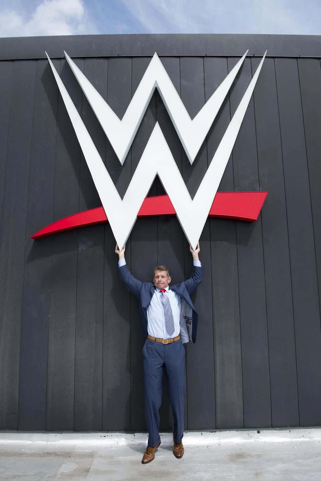 WWE Logo 桌布 壁紙 — 一名身穿西裝的男子在明亮的藍天下，自豪地舉起一個巨大的WWE標誌，背景是深灰色的牆壁。