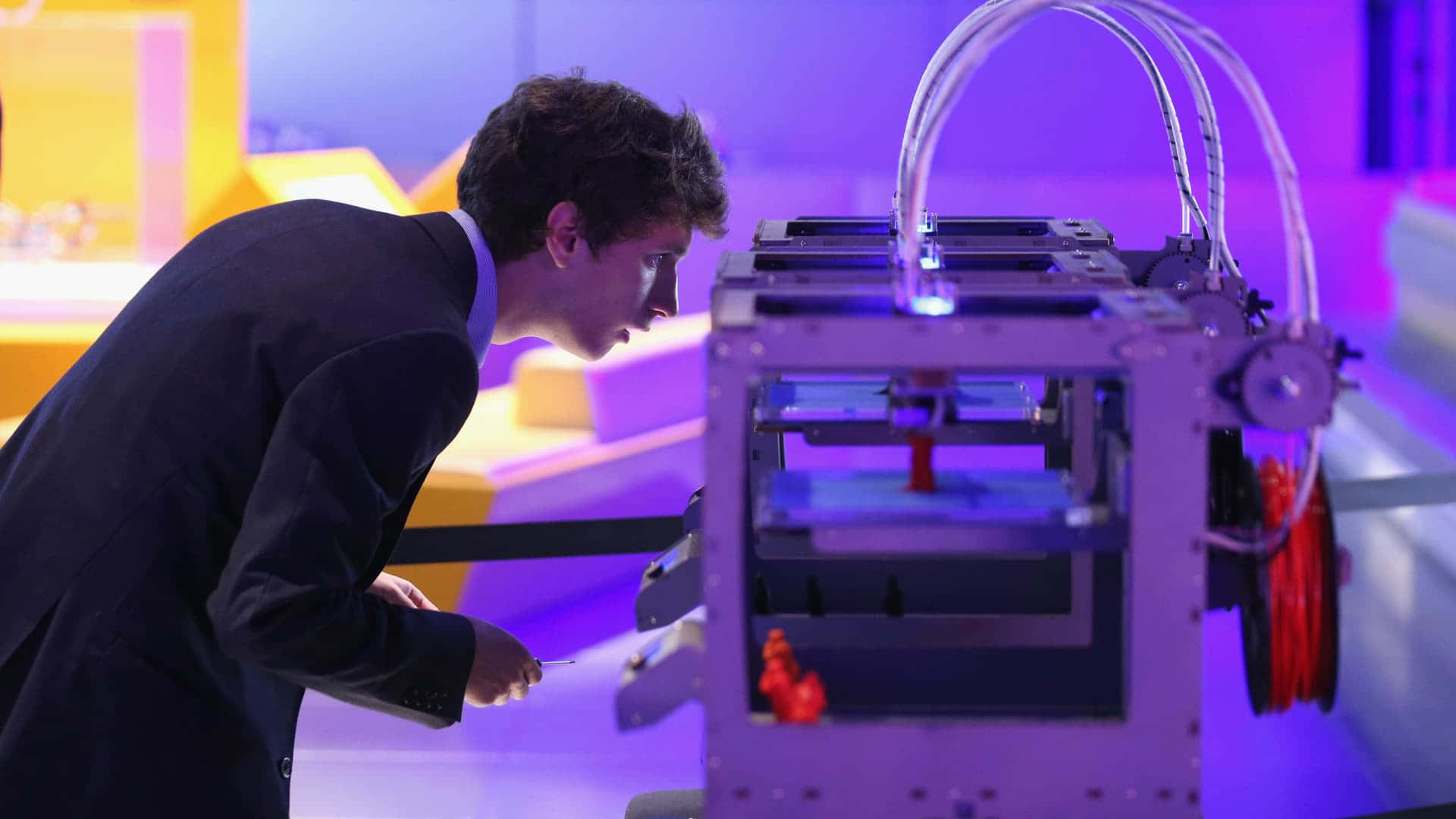 3d Printer 桌布 壁紙 — 一名身穿西裝的男子在現代科技展覽中，專注地俯身靠近一部散發著紫色和藍色光芒的3D打印機。