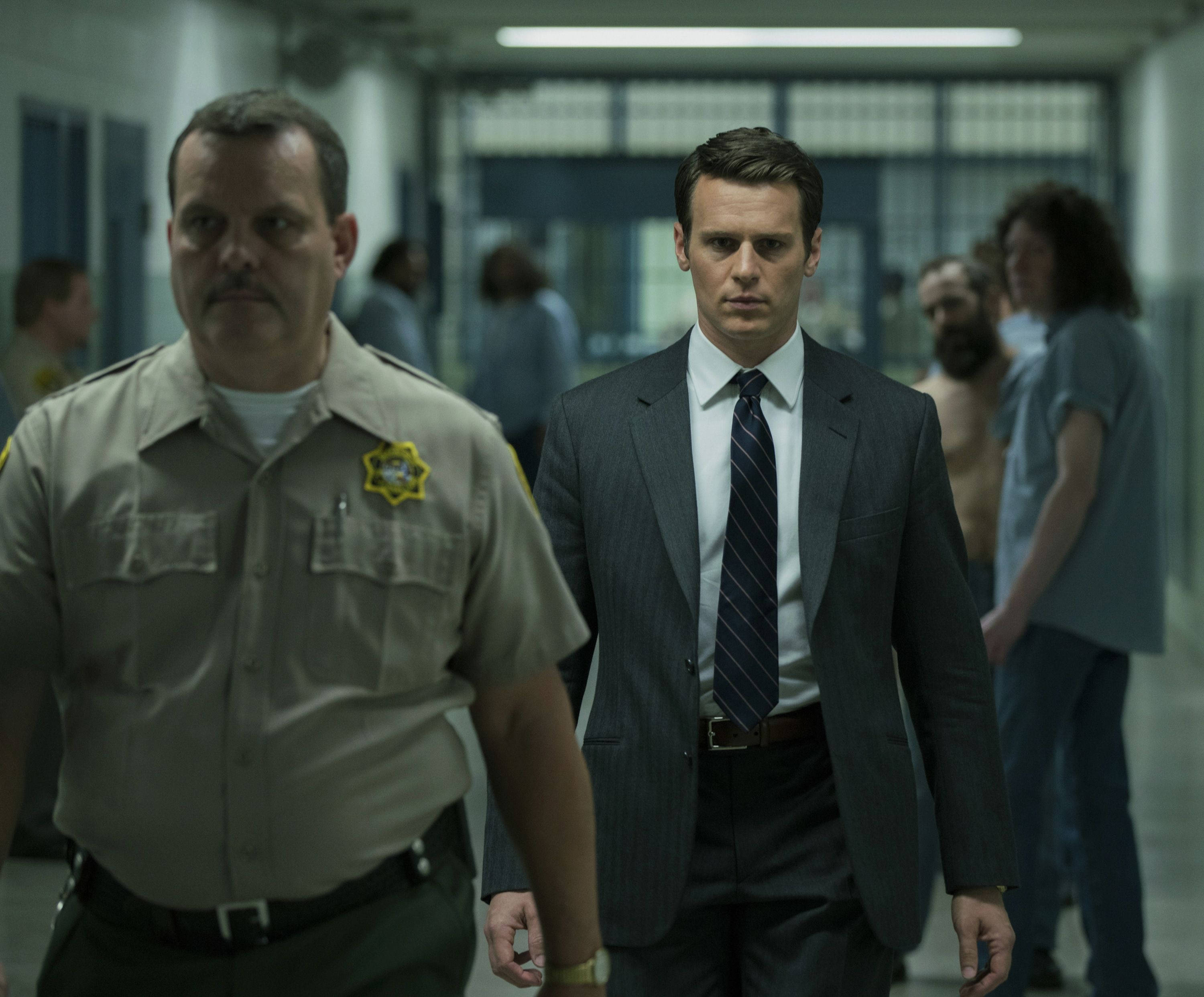 Jonathan Groff Stars In Netflix Mindhunter Series 壁紙 — 一名身穿西裝的男子在監獄走廊行走，兩旁有警衛和背景中的囚犯。