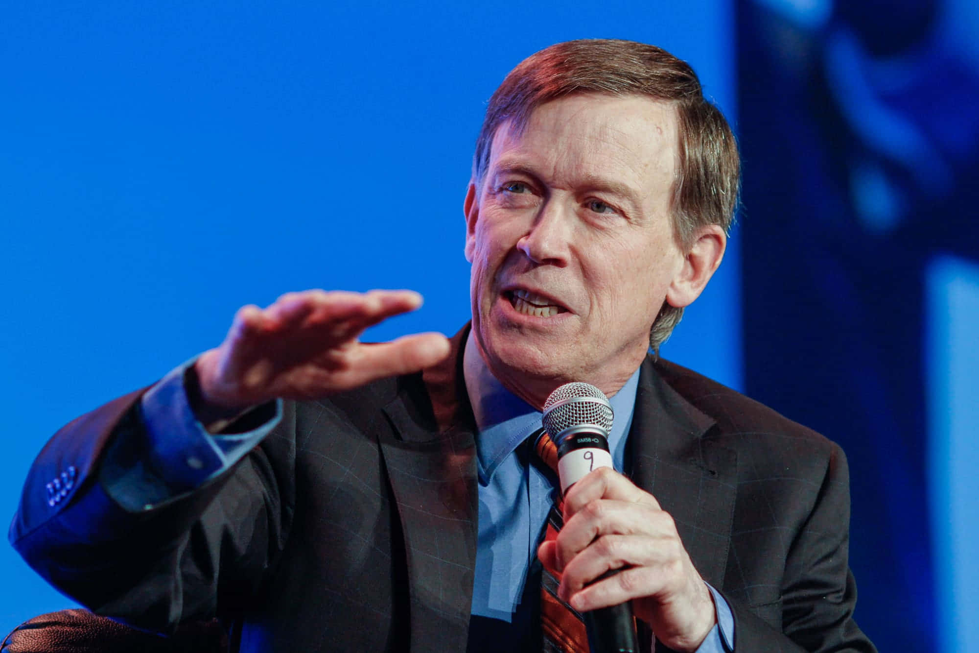Blue Aesthetic John Hickenlooper With Microphone 壁紙 — 一名身穿西裝的男子在純藍色背景前對著麥克風說話並做手勢。