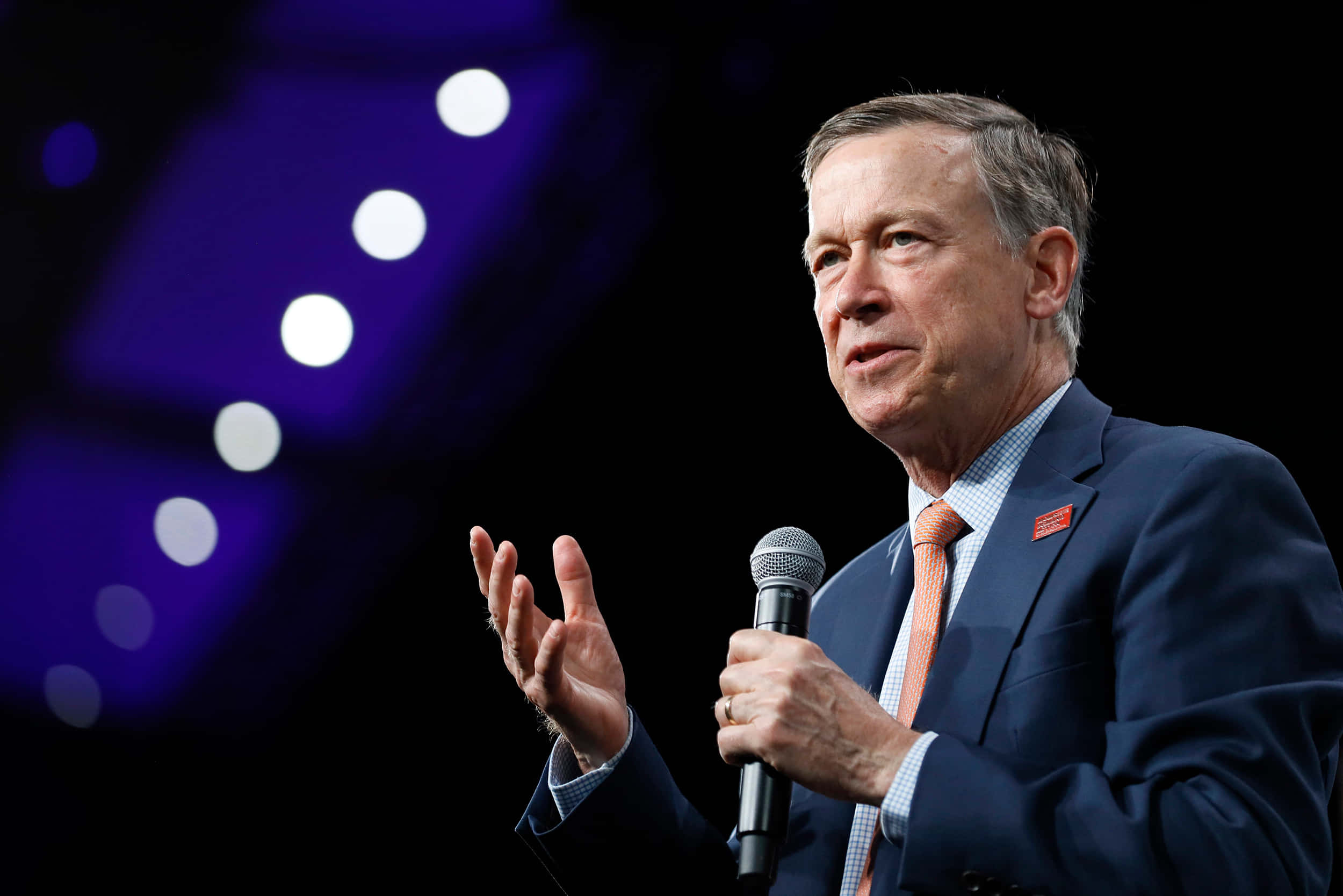 John Hickenlooper addressing public 壁紙 — 一名身穿西裝的男子在黑暗的舞台上對著麥克風說話，舞台上有紫色燈光和散景效果。