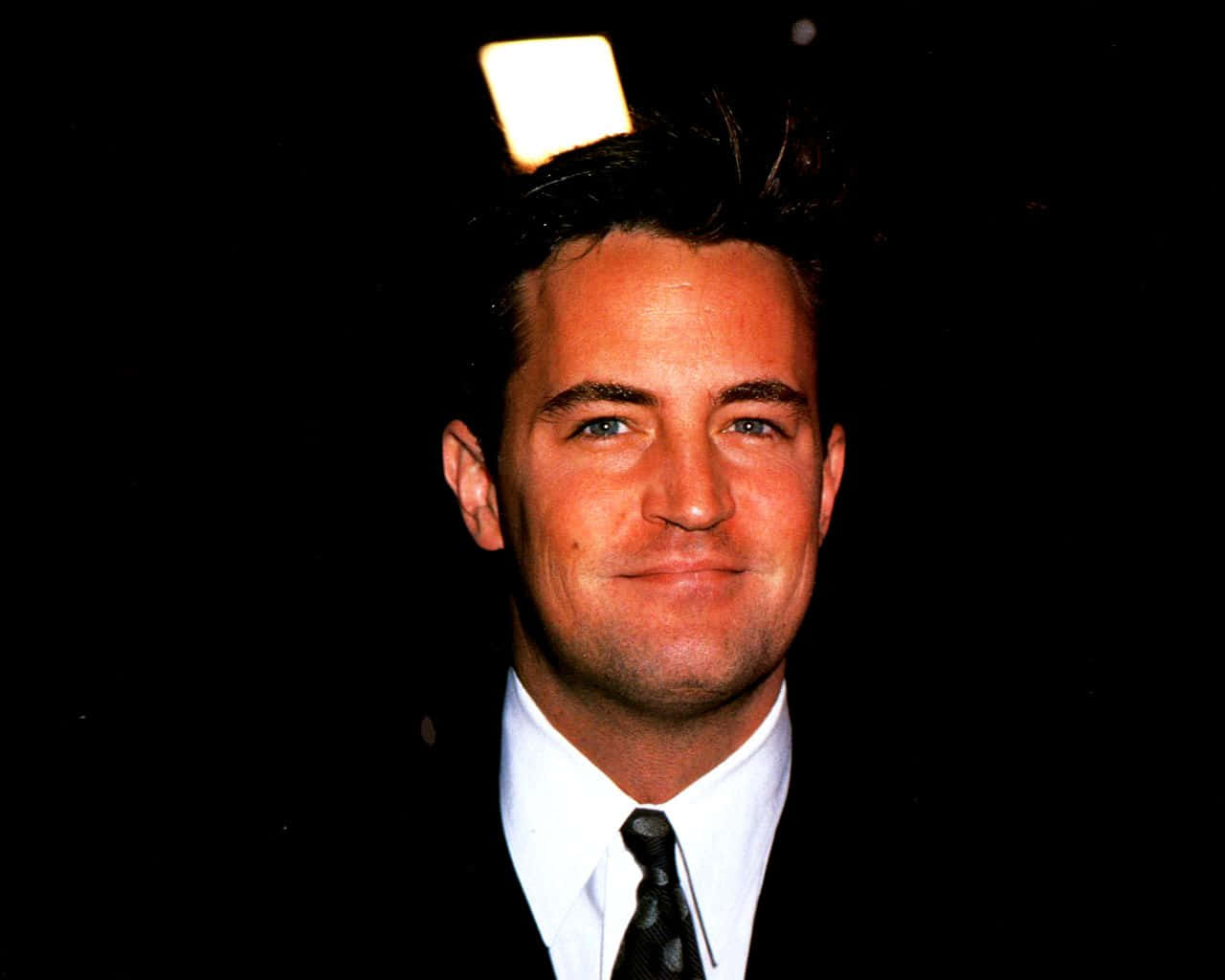 matthew perry 桌布 壁紙 — 一名身穿西裝的男子在黑色背景前微笑，頭頂上方有一個發光的正方形。