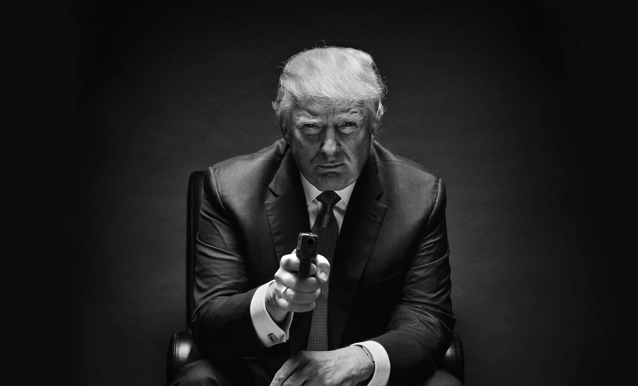 trump 桌布 壁紙 — 一名身穿西裝的男子坐在椅子上，手持手槍指向前方，背景為深色。