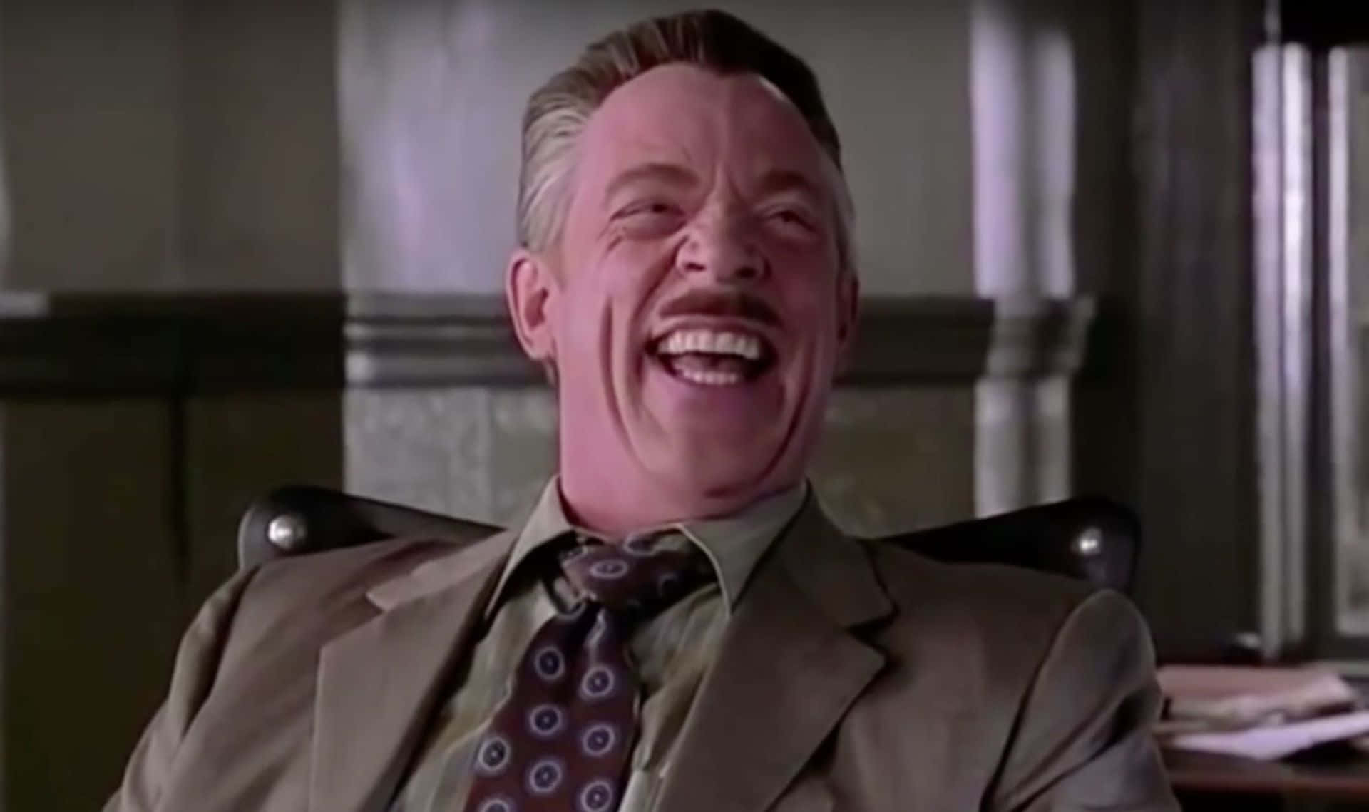 J Jonah Jameson 桌布 壁紙 — 一名身穿西裝的男子坐在辦公椅上，戴著圖案領帶，開懷大笑。