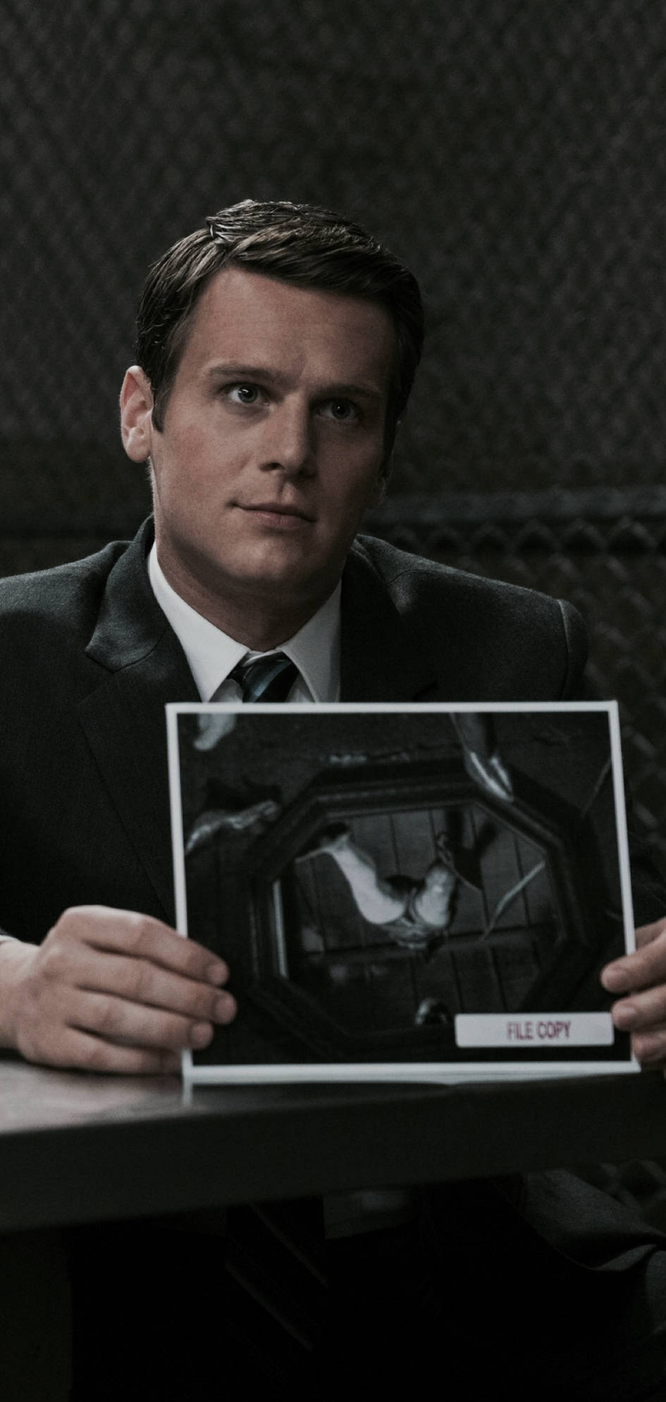 Jonathan Groff Mindhunter Monster Exposé 壁紙 — 一名身穿西裝的男子手持一張標有「檔案副本」的照片，照片中顯示一隻鳥在籠子裡，背景是深色的鐵絲網。