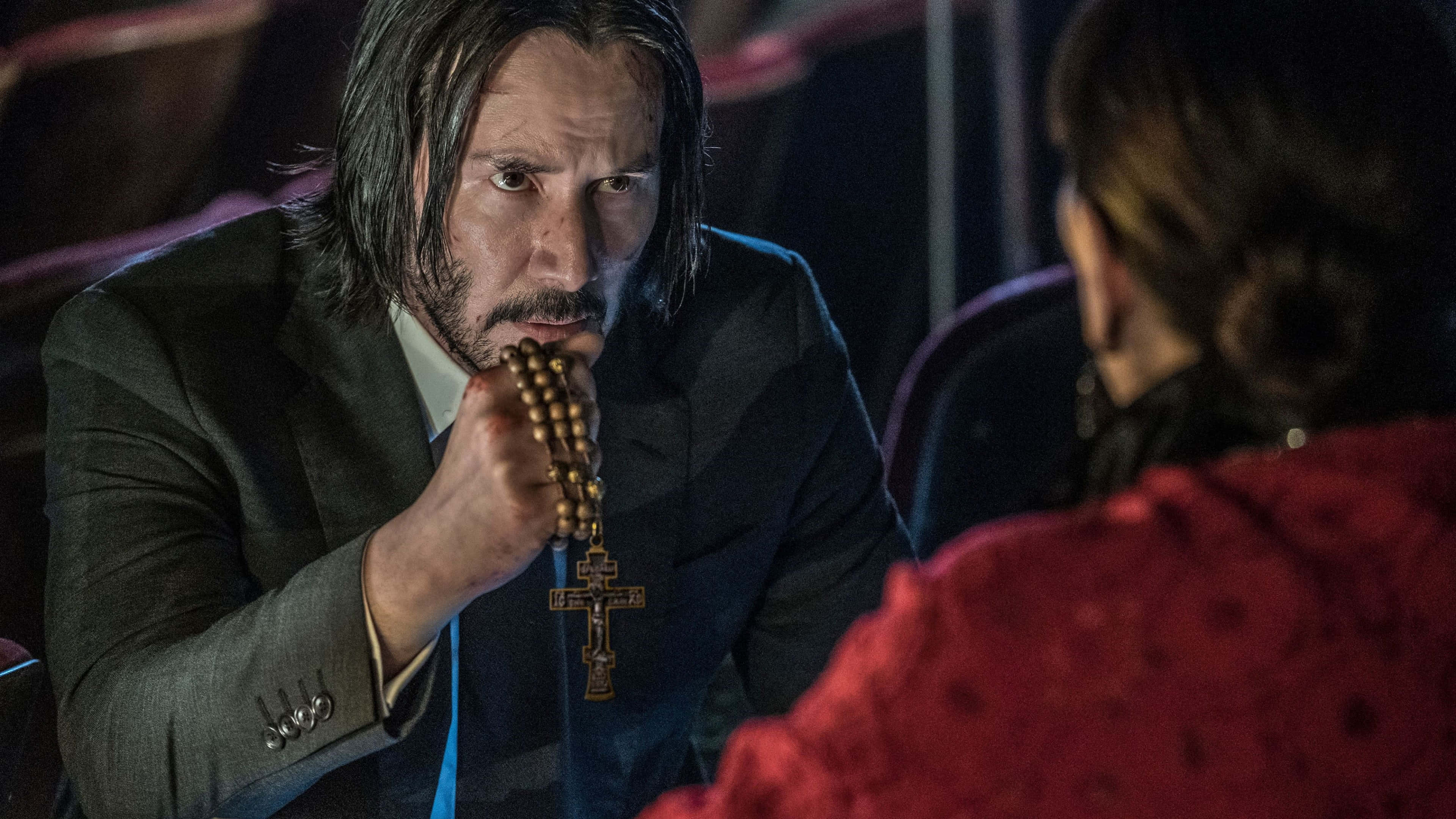 Keanu Reeves 桌布 壁紙 — 一名身穿西裝的男子手持玫瑰經念珠，在燈光昏暗的劇院中凝視著另一個人。