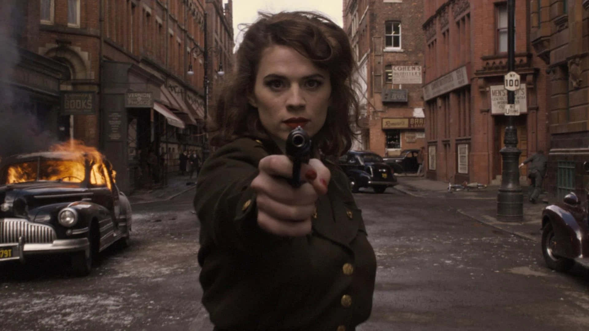 Peggy Carter 桌布 壁紙 — 一名身穿軍裝外套的女性，在骯髒的城市街道上，直接用手槍瞄準觀眾，附近有一輛燃燒的復古汽車。