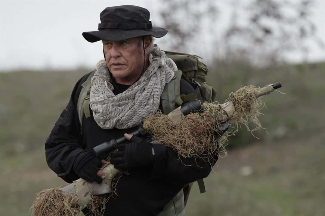 Tom Berenger 桌布 壁紙 — 一名身穿迷彩服和奔尼帽的士兵在荒蕪的田野中手持一把用草偽裝的步槍。