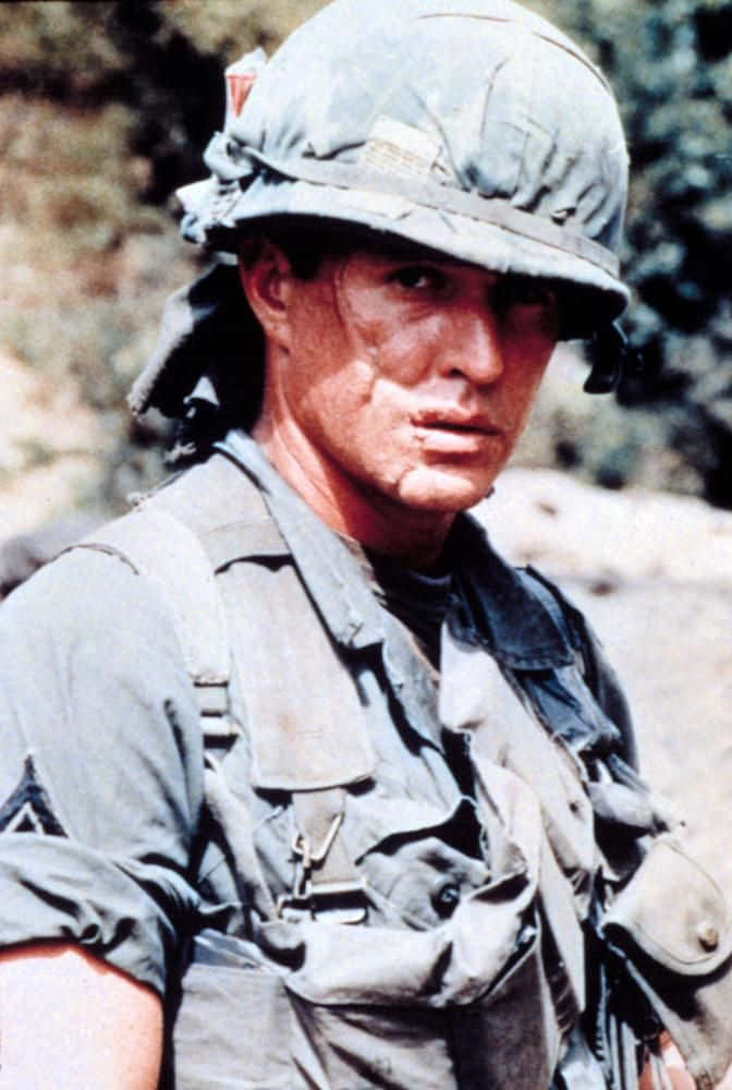 Tom Berenger 桌布 壁紙 — 一名身穿迷彩服和頭盔的疲憊士兵，在模糊的自然背景下，以曬傷的臉龐專注地凝視。