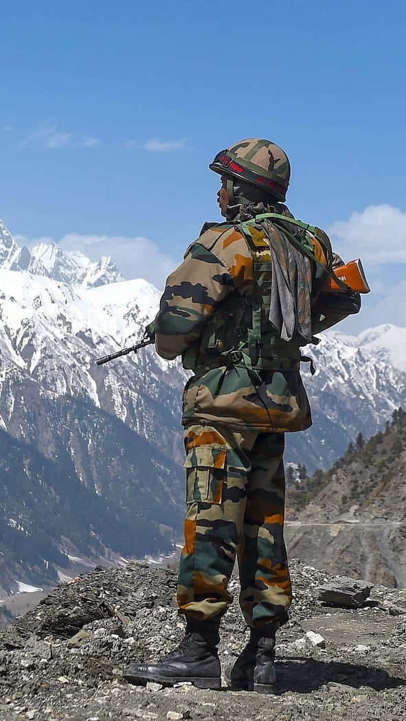 Indian Army 桌布 壁紙 — 一名身穿迷彩服的士兵在岩石山脊上站崗，俯瞰著晴朗藍天下白雪覆蓋的山峰。