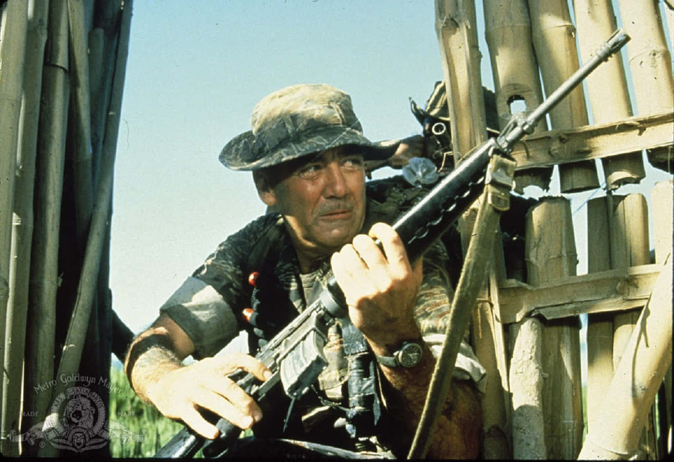 R. Lee Ermey 桌布 壁紙 — 一名身穿迷彩裝備的士兵在明亮的日光下，從竹製路障後方專注地凝視，手持突擊步槍。