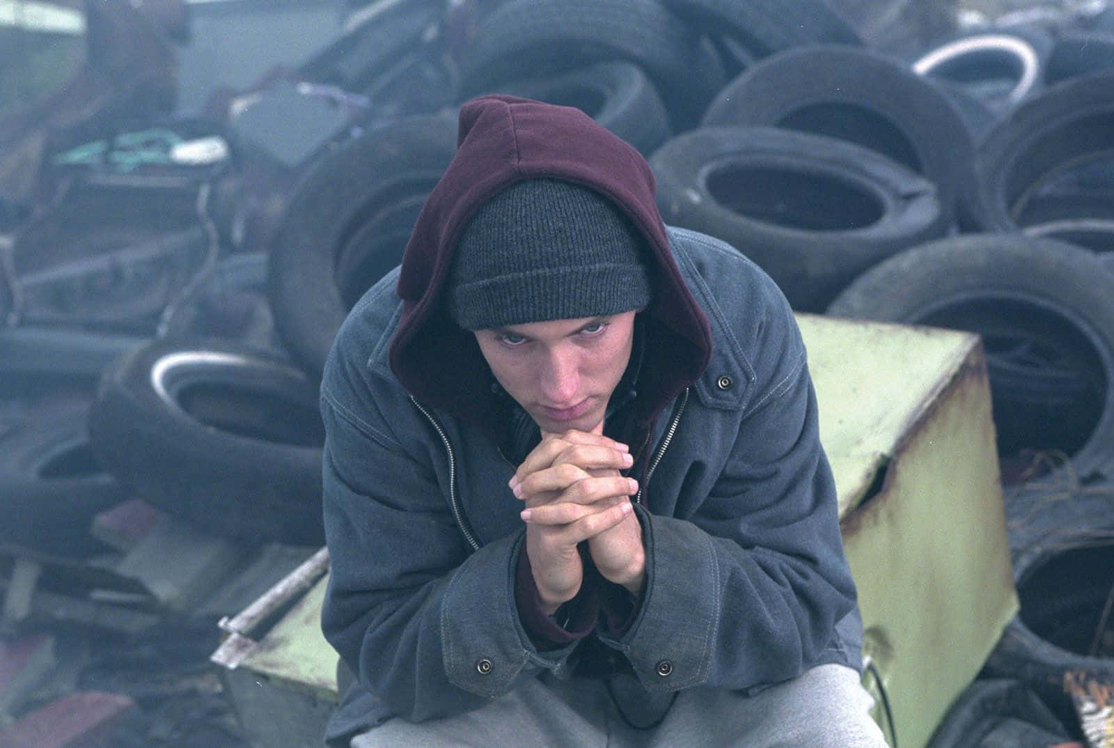 Eminem 桌布 壁紙 — 一名身穿連帽衛衣的年輕男子在廢棄輪胎中莊嚴跪下，雙手合十，靜默沉思。