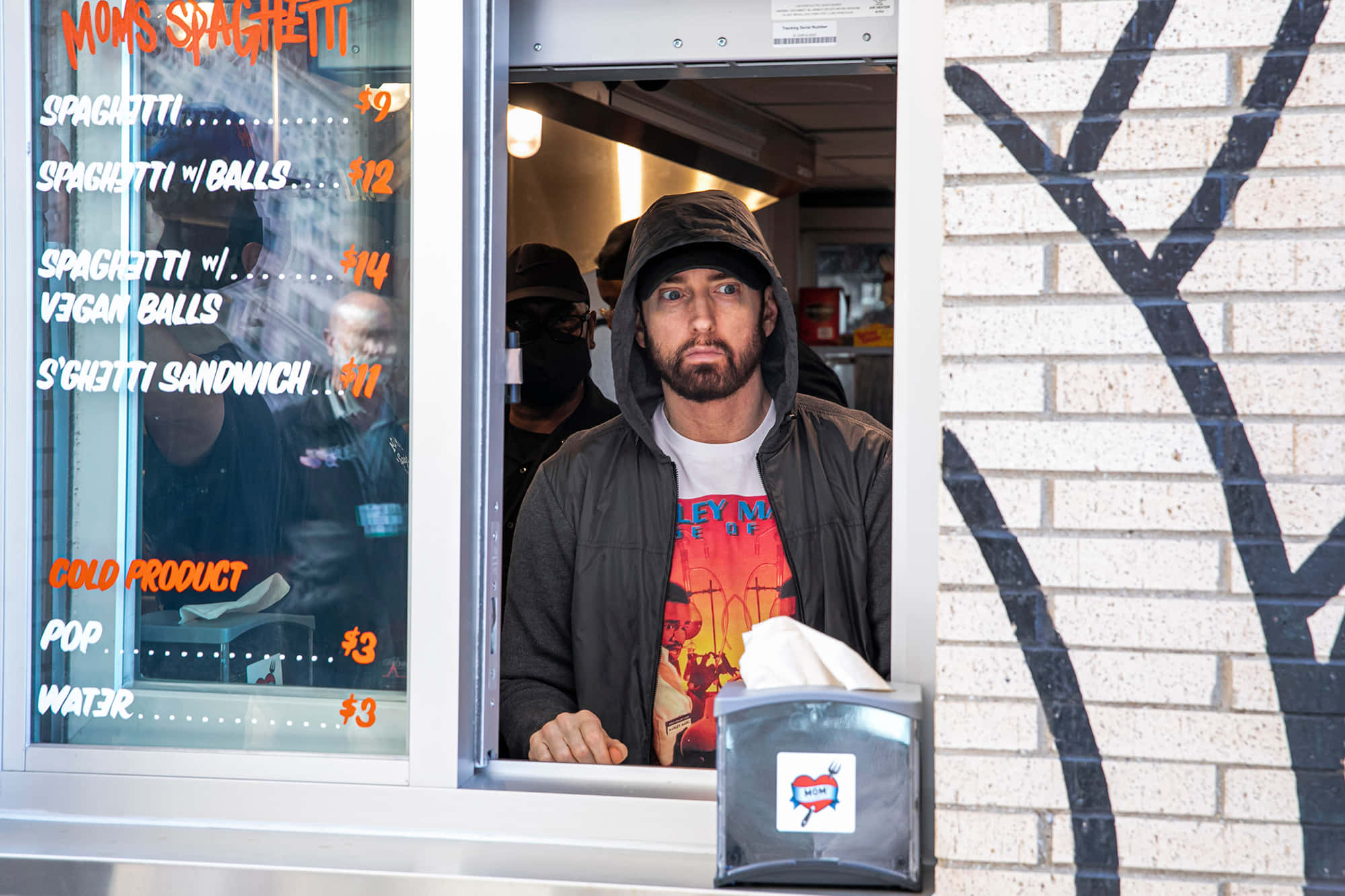 Eminem 桌布 壁紙 — 一名身穿連帽衫的男子站在一輛餐車窗前，菜單上列出意大利麵和純素丸子。