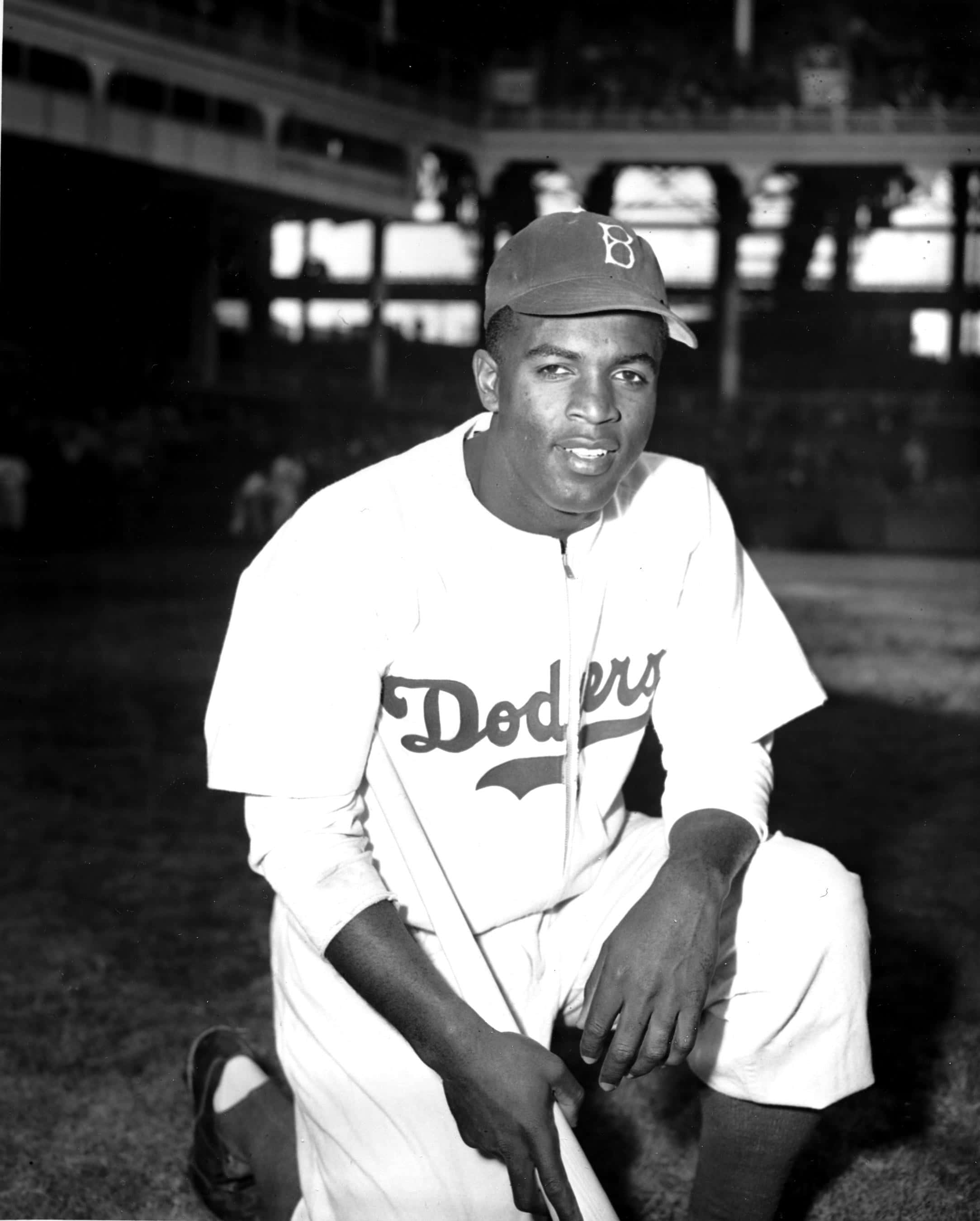 Jackie Robinson 桌布 壁紙 — 一名身穿道奇隊制服、面帶微笑的棒球運動員在夜間球場燈光下單膝跪地，手持球棒。