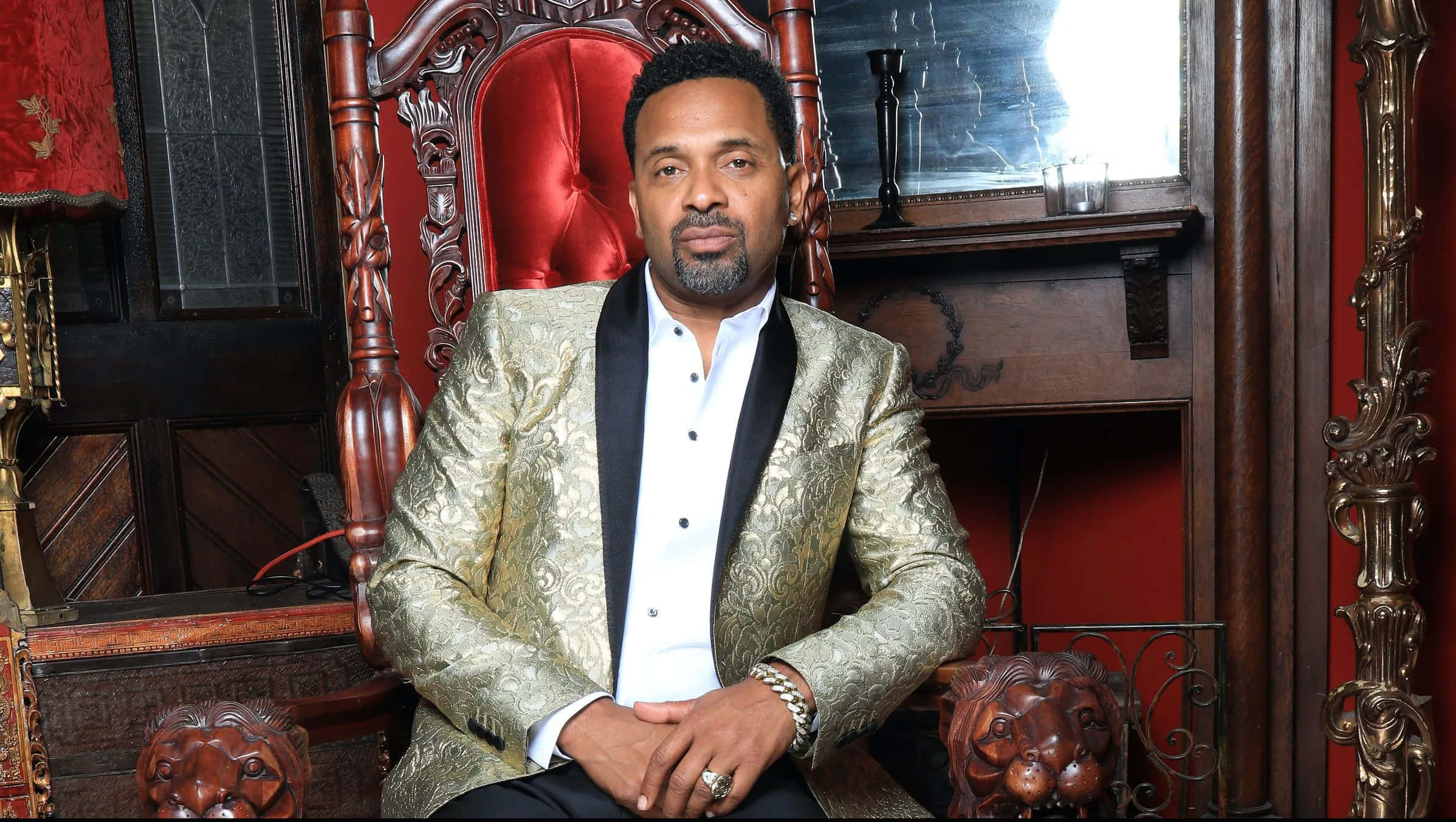 Mike Epps 桌布 壁紙 — 一名身穿金色錦緞外套的男子，在裝飾華麗的房間裡，威嚴地坐在一張華麗的紅色寶座椅上。