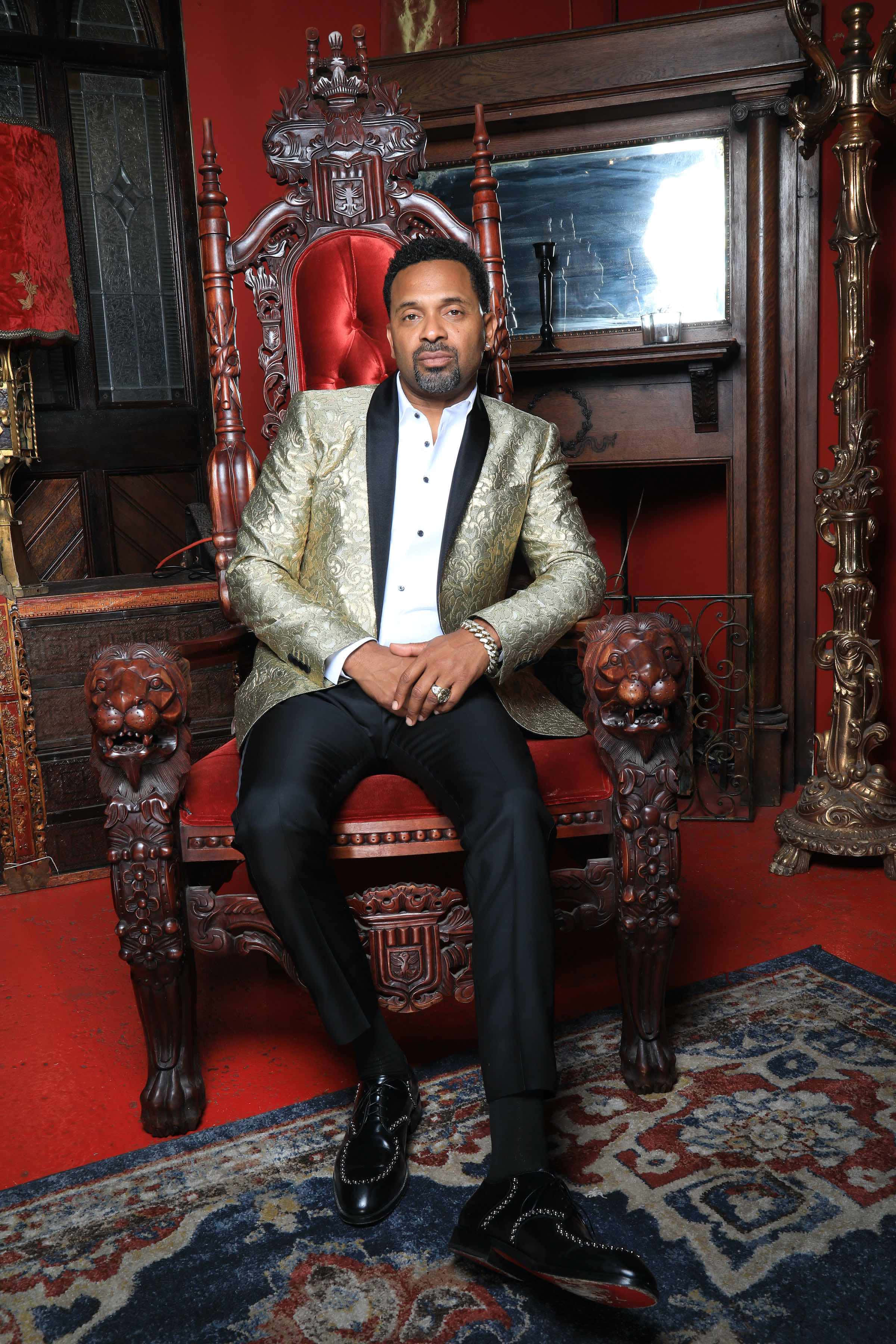 Mike Epps 桌布 壁紙 — 一名身穿金色錦緞外套的男子，威嚴地坐在一張華麗的紅色寶座椅上，房間裝飾華麗，有深紅色牆壁和圖案地毯。