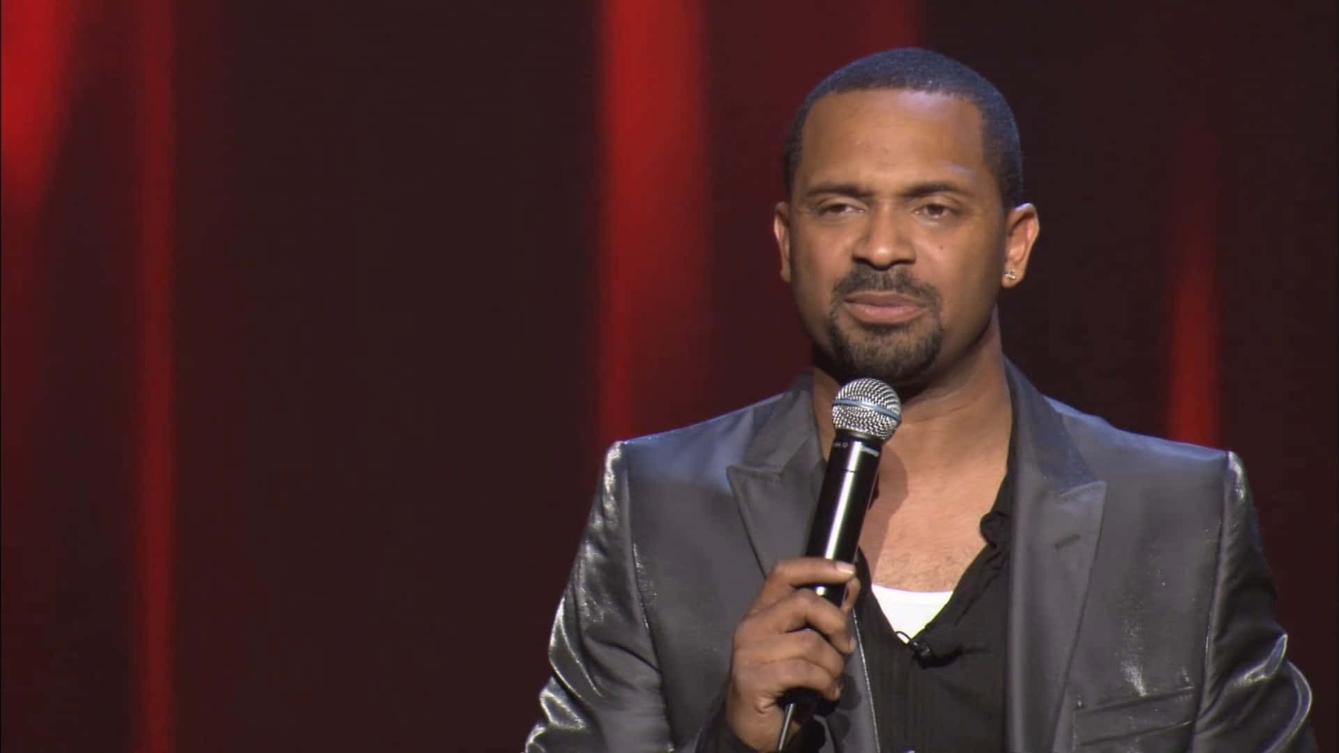 Mike Epps 桌布 壁紙 — 一名身穿閃亮灰色西裝的男子，在深紅色窗簾背景下手持麥克風。