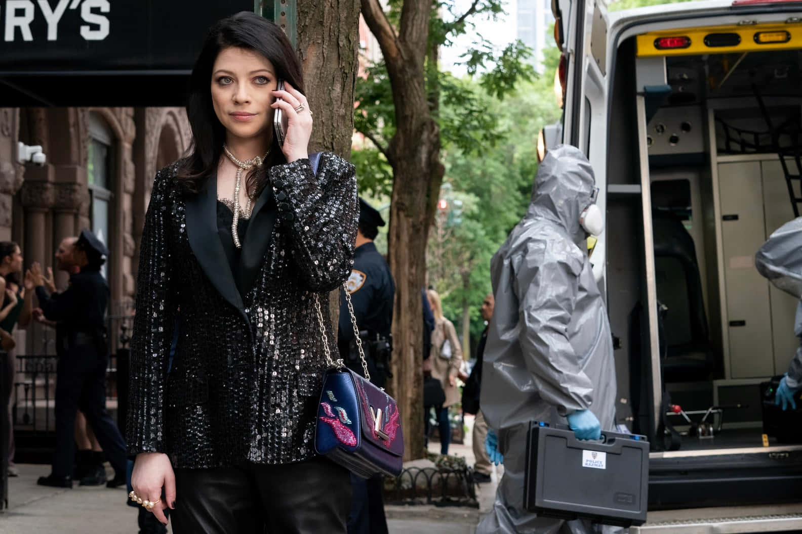 michelle trachtenberg 桌布 壁紙 — 一名身穿閃亮黑色西裝外套的女子正在講電話，而穿著防護服的鑑證小組在救護車旁的城市街道上工作。