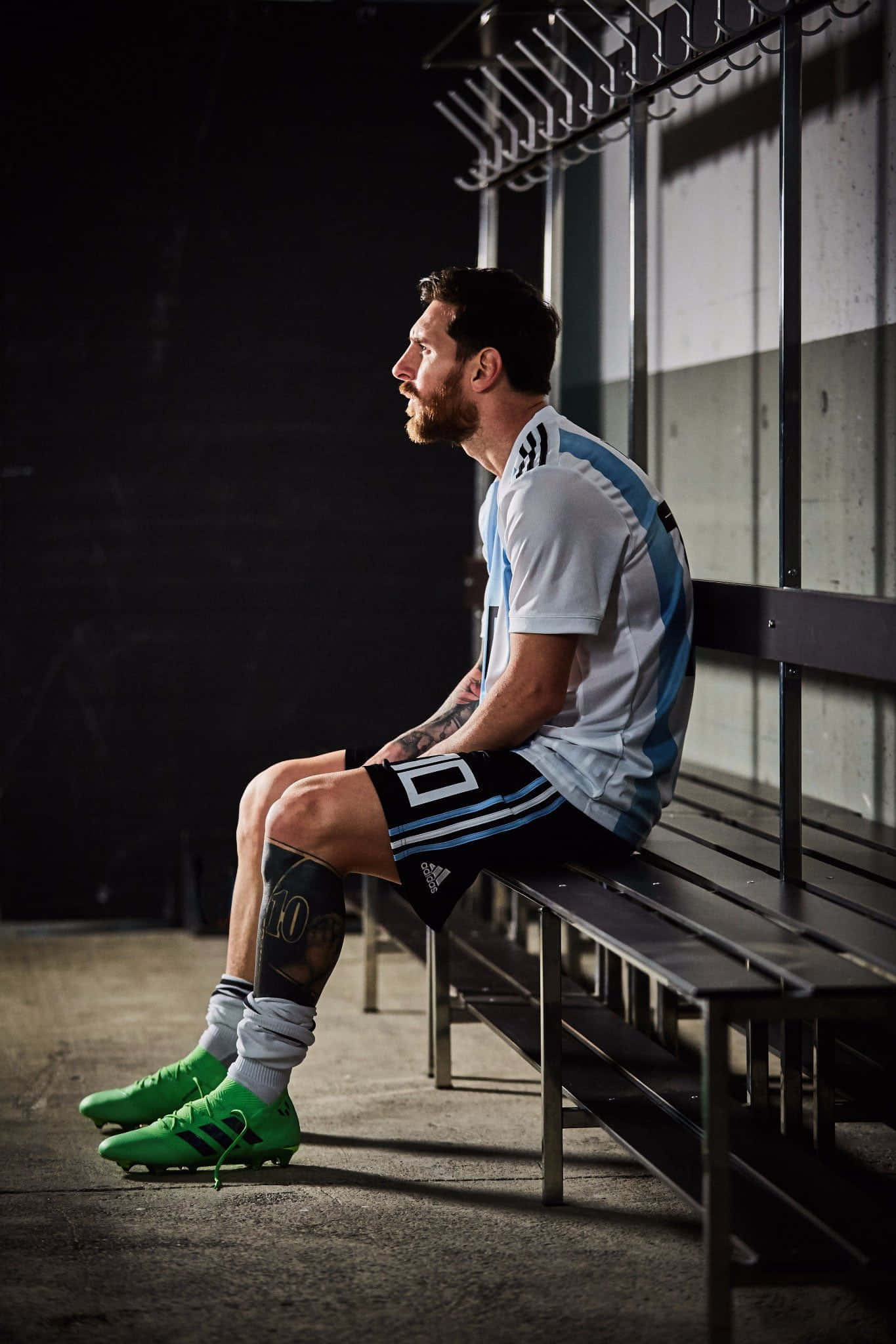 Messi 2022 桌布 壁紙 — 一名身穿阿根廷藍白球衣的足球員，在燈光昏暗的更衣室裡，若有所思地坐在金屬長凳上，穿著鮮綠色球鞋。