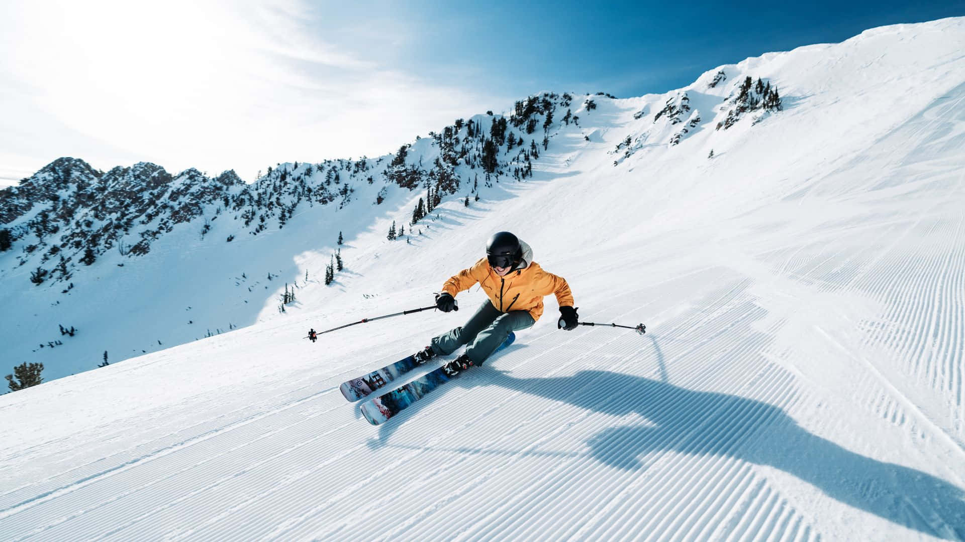 Skiing 桌布 壁紙 — 一名身穿鮮橙色外套的滑雪者，在晴朗藍天下，以岩石山峰為背景，沿著修整過的雪坡滑行而下。