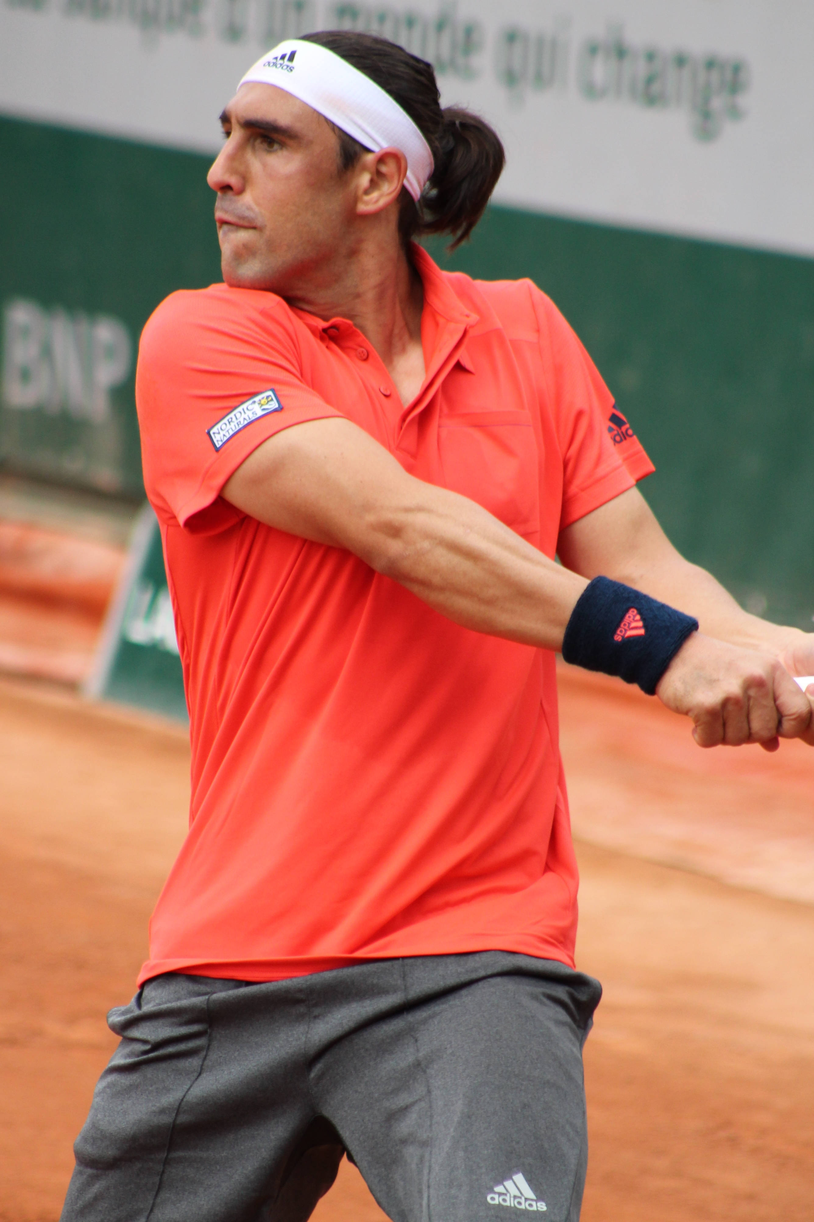 Marcos Baghdatis Orange Adidas Top 壁紙 — 一名身穿鮮橙色襯衫的男網球運動員在泥地球場上專注地揮拍。