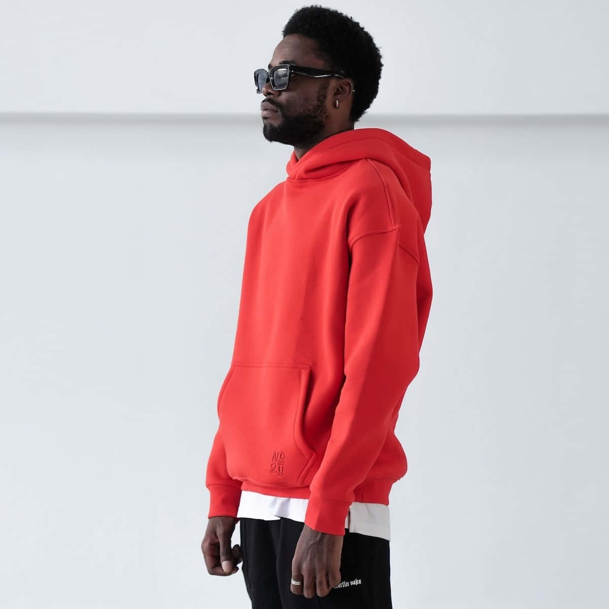 Red Hoodie 桌布 壁紙 — 一名身穿鮮紅色連帽衛衣和黑色太陽眼鏡的男子，在極簡白色背景前側面站立。