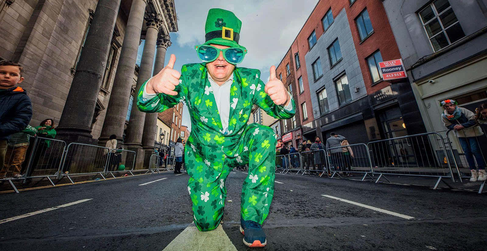 St Patrick's Day 桌布 壁紙 — 一名身穿鮮綠色三葉草西裝和帽子的男子在聖帕特里克節遊行期間跑過城市街道，豎起兩個大拇指。