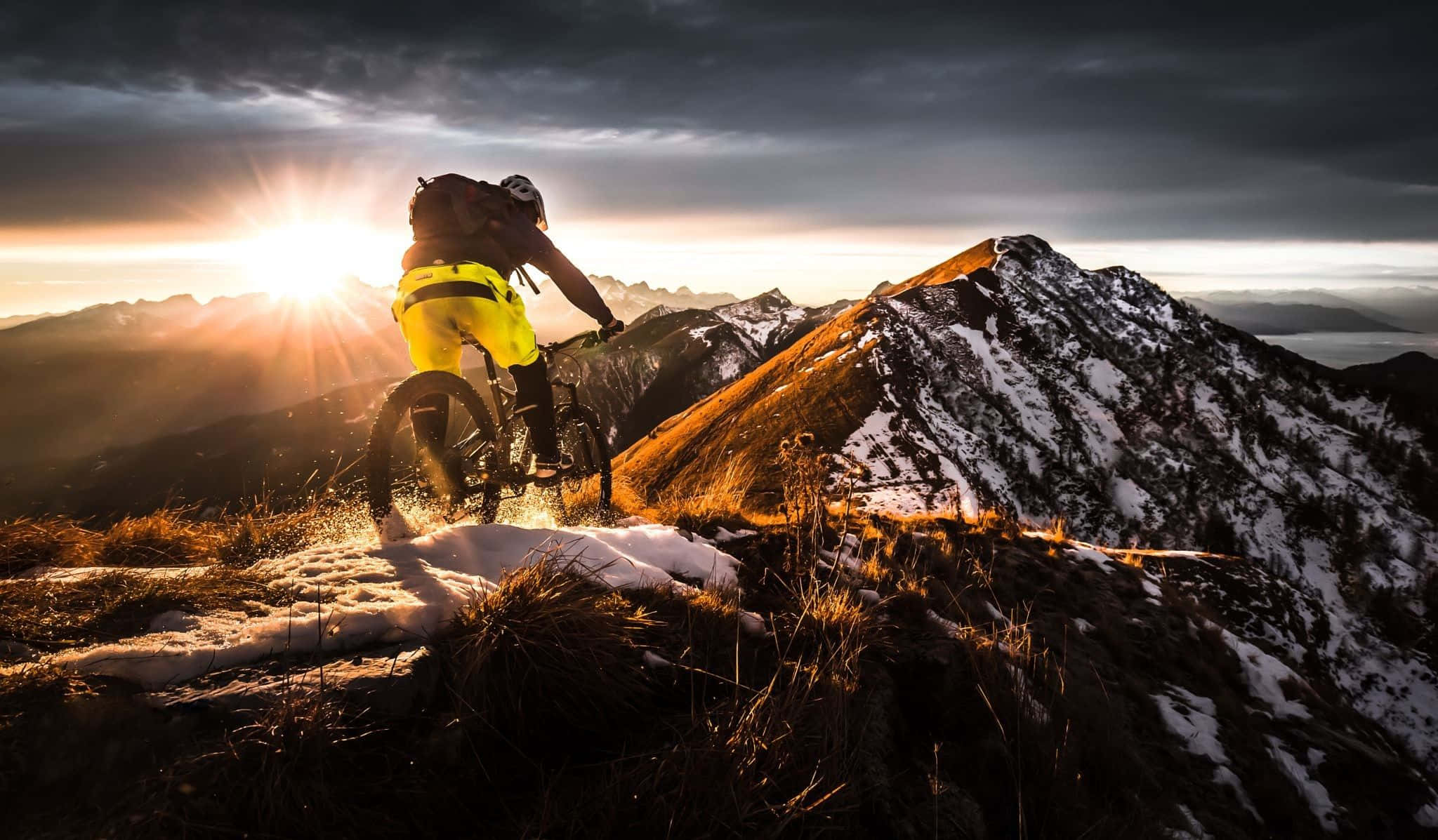 Mtb 桌布 壁紙 — 一名身穿鮮黃色裝備的山地車手在崎嶇的山峰上穿過雪地和草地，陽光從暴風雲中迸發而出。