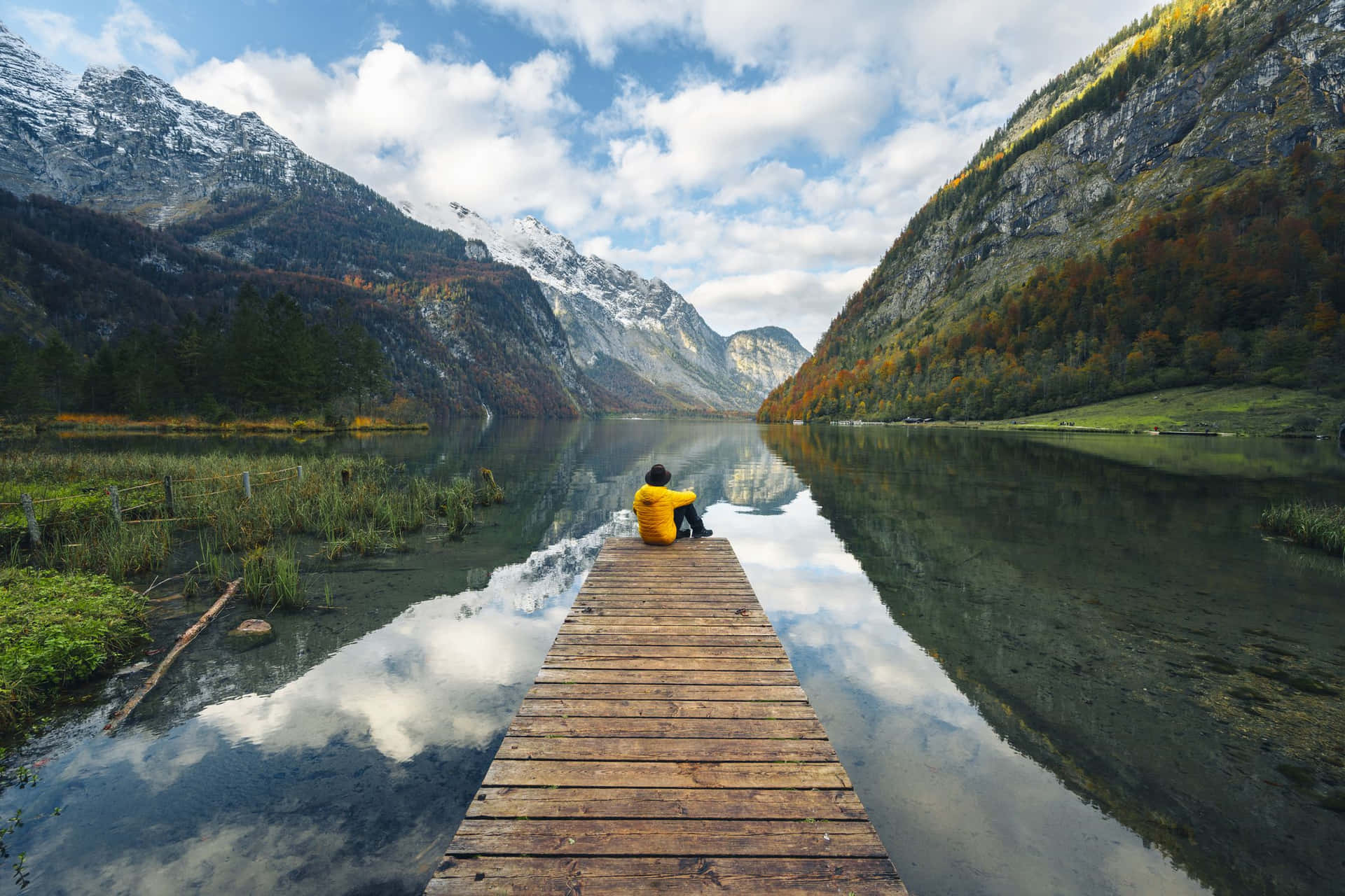 Nature Scenery 桌布 壁紙 — 一名身穿黃色外套的人獨自坐在延伸至寧靜山湖的木製碼頭上，周圍是秋日森林和積雪的山峰，天空部分多雲。