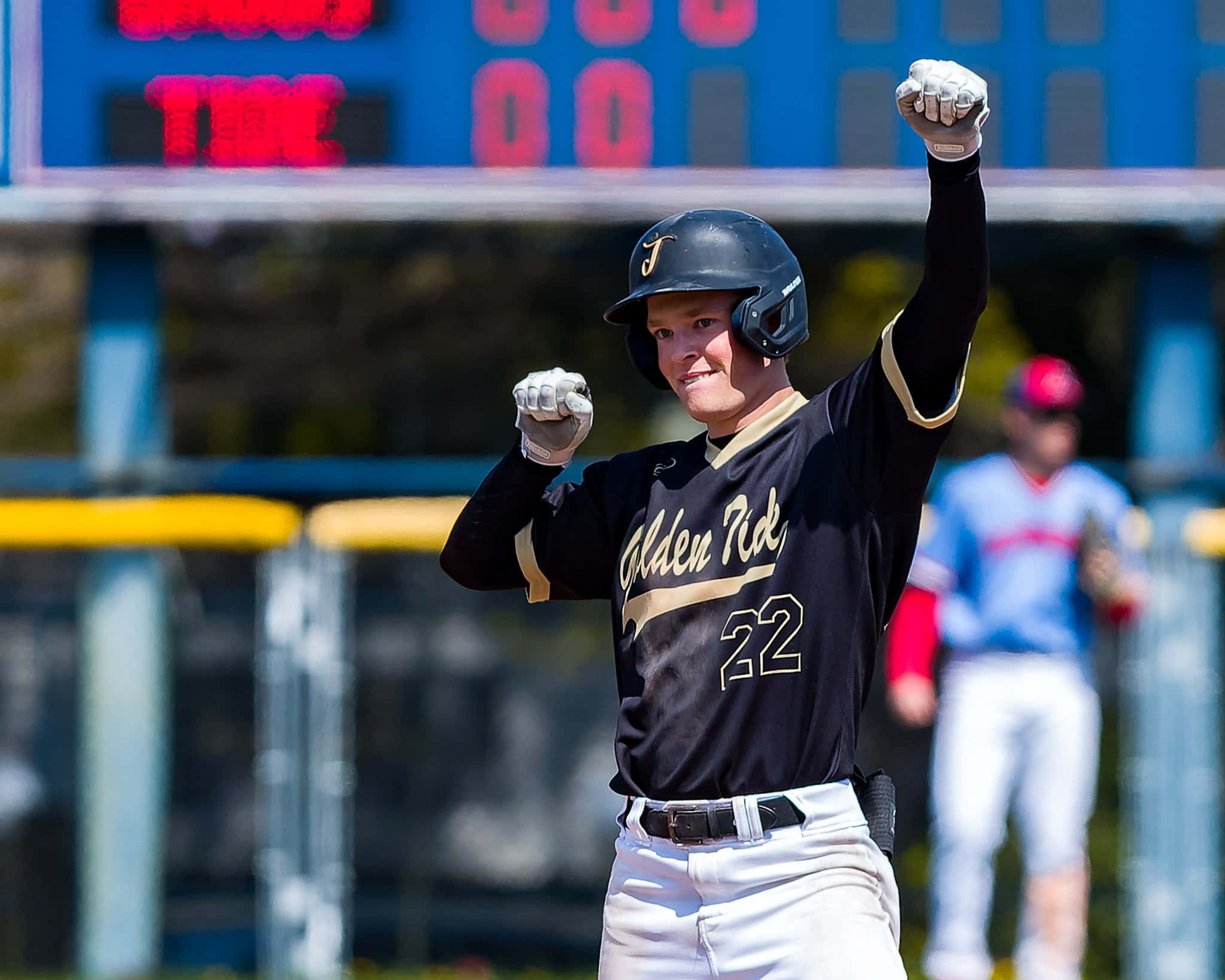 College Baseball 桌布 壁紙 — 一名身穿黑色「Golden Rick」球衣的棒球運動員在球場明亮的日光下高舉雙拳慶祝勝利。