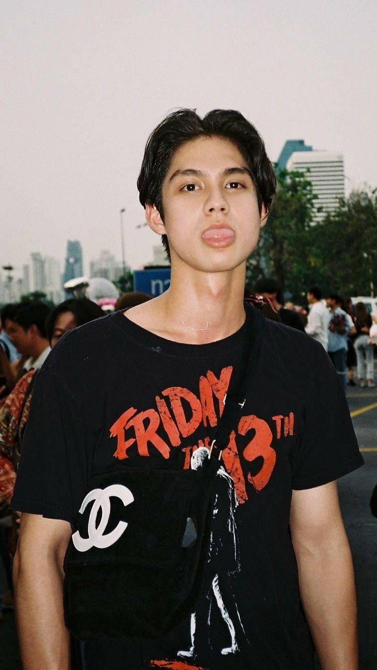 Bright Vachirawit 桌布 壁紙 — 一名身穿黑色Friday the 13th T恤的年輕男子，在擠滿人群的城市街道上嘟起嘴唇，背景是高樓大廈。